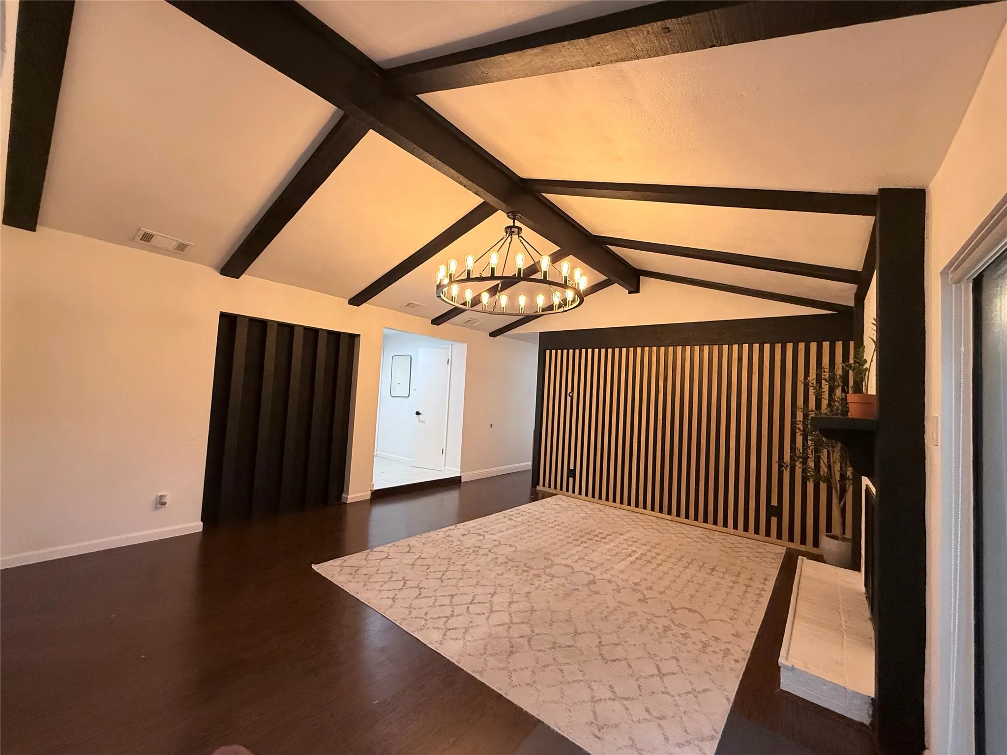 Vaulted ceiling with exposed beams, wood-finish flooring, fireplace with white surround, and decorative slat wall