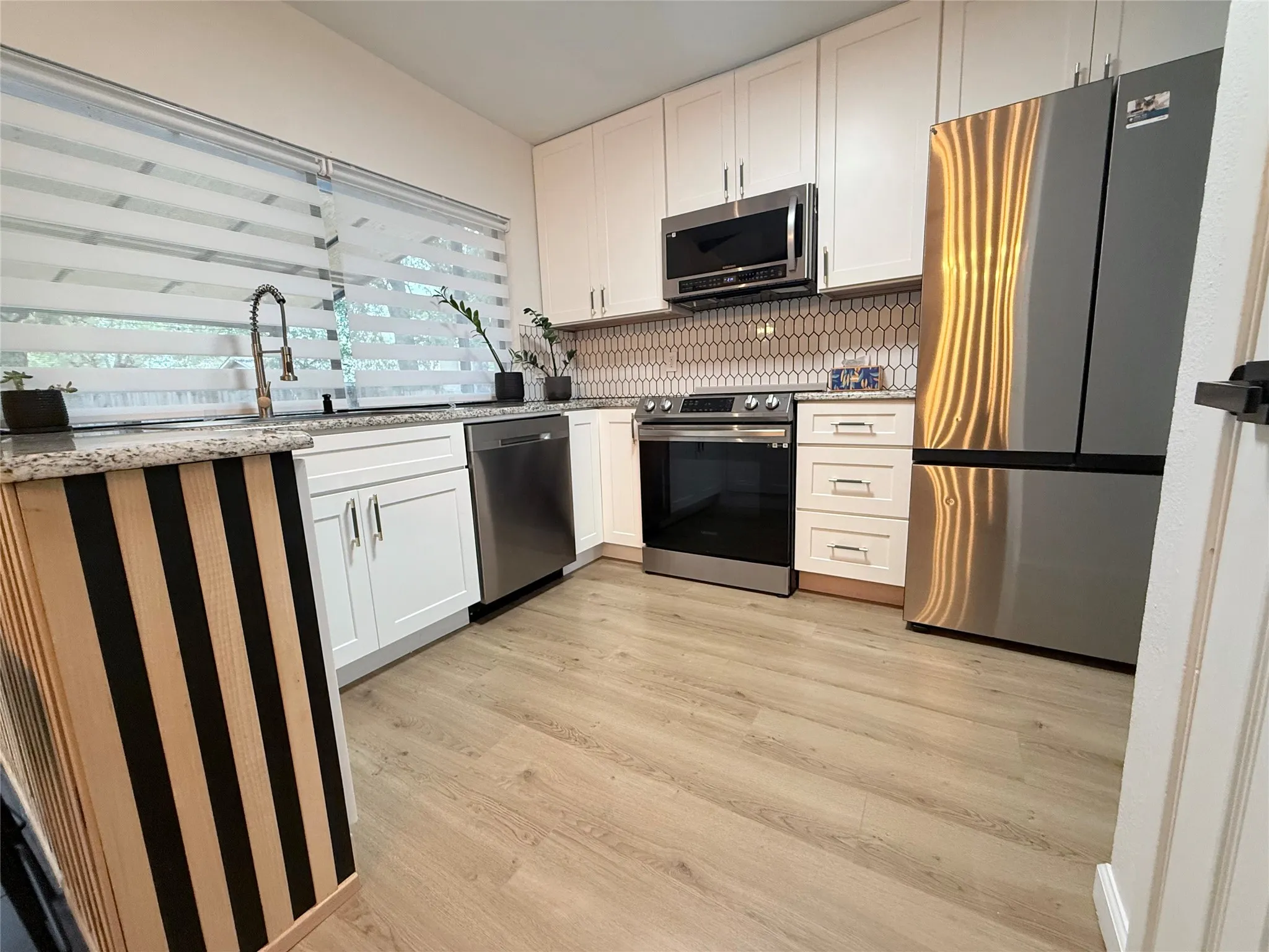 Wood-finish flooring, white cabinetry, stainless steel appliances, speckled countertops, and an elongated hexagon tile backsplash