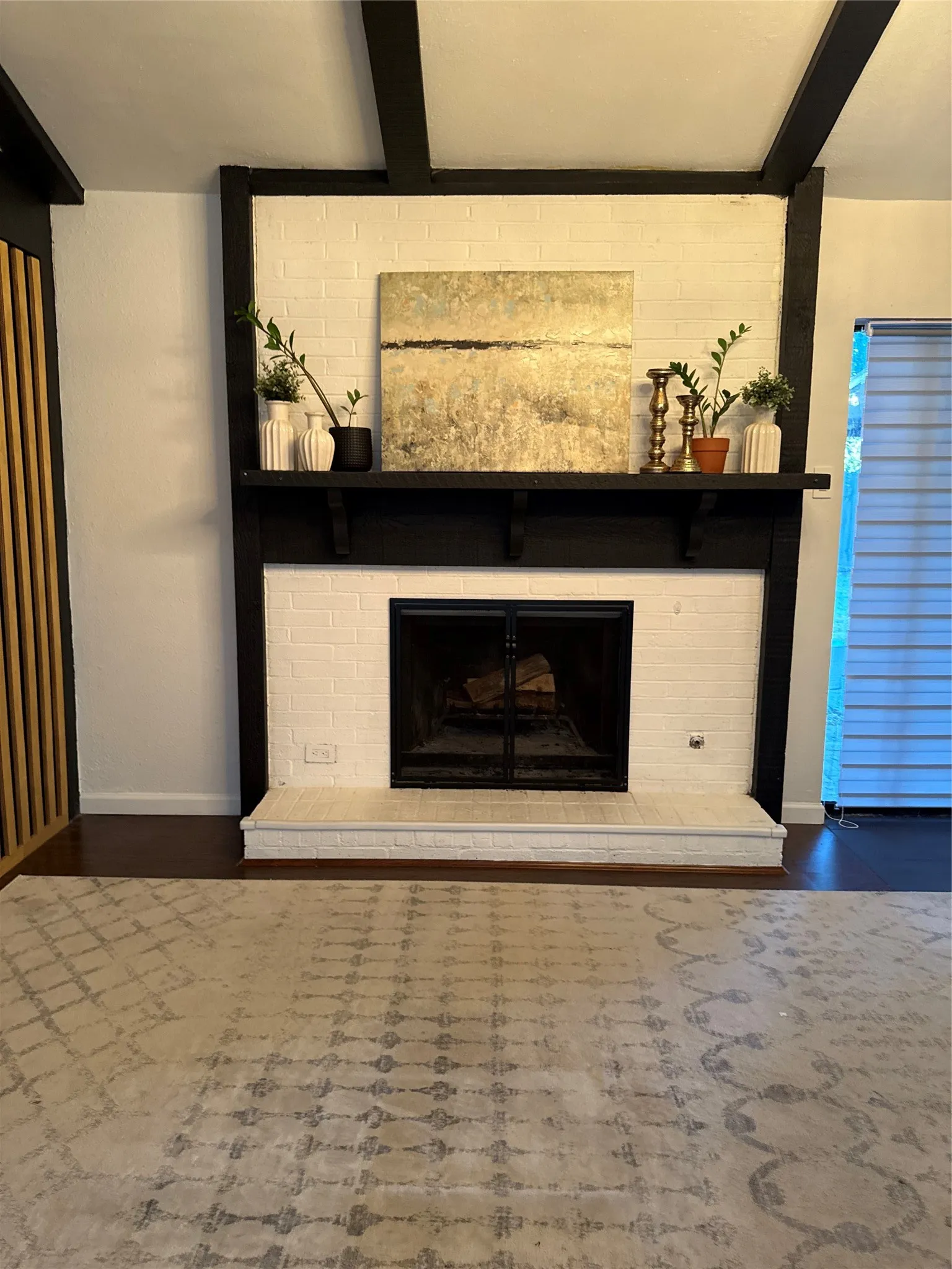 White brick fireplace with a black mantel and hearth, dark wood-finish flooring, and black ceiling beams