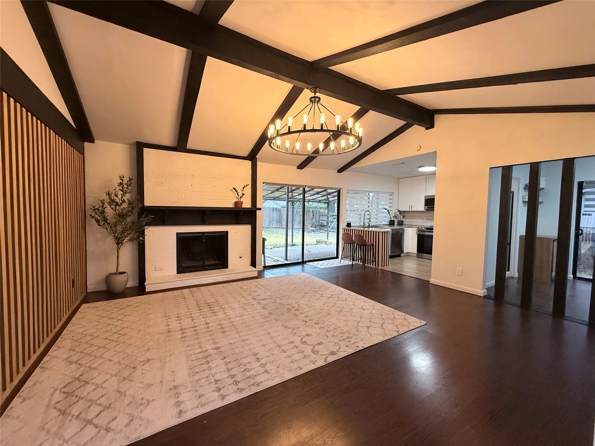 Vaulted ceiling with exposed dark beams, dark wood-finish flooring, white brick fireplace with dark mantel, wood slat accent wall, and sliding glass doors