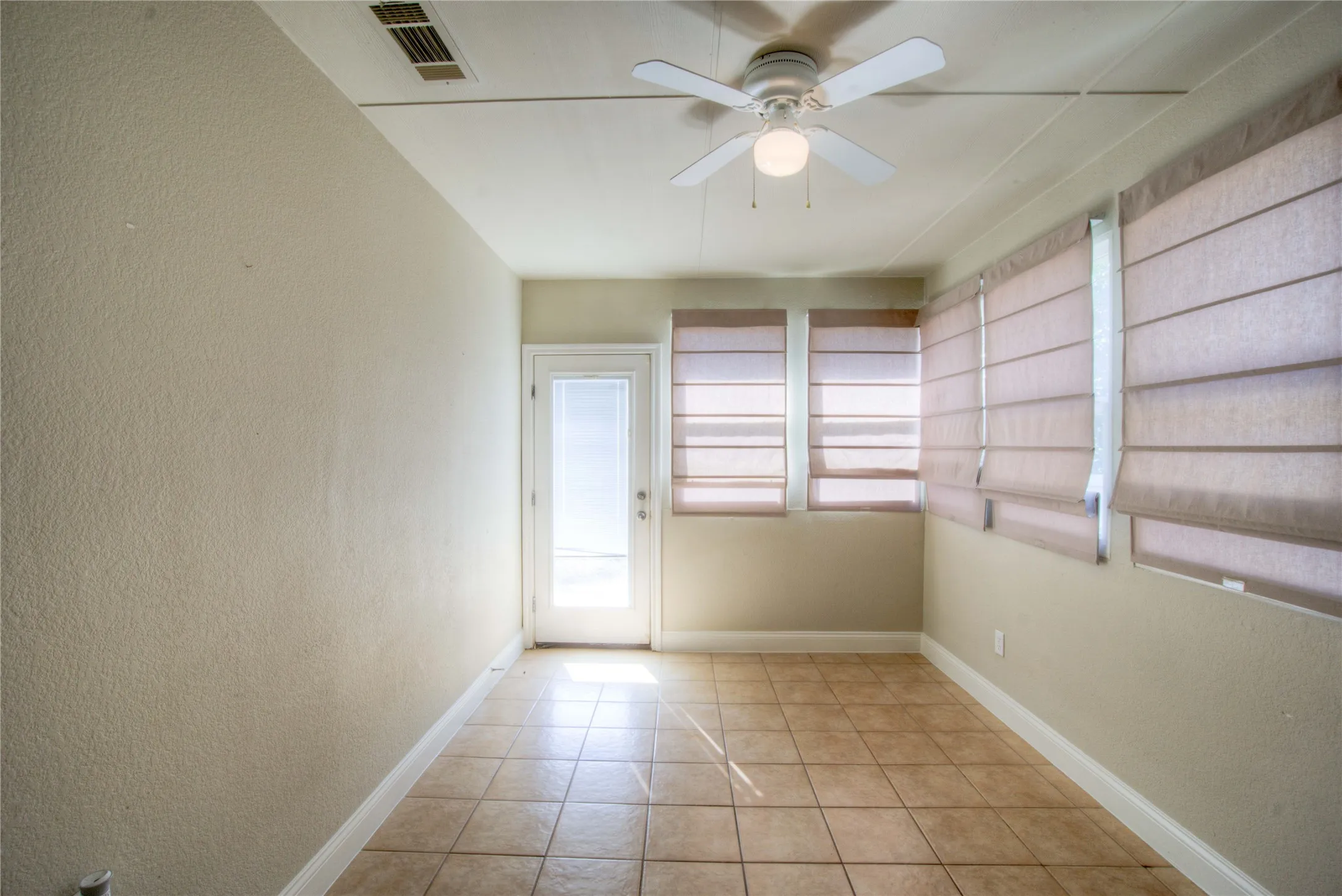 Tile flooring, ceiling fan with integrated lighting, glass-paneled door, multiple windows with Roman shades, and textured walls