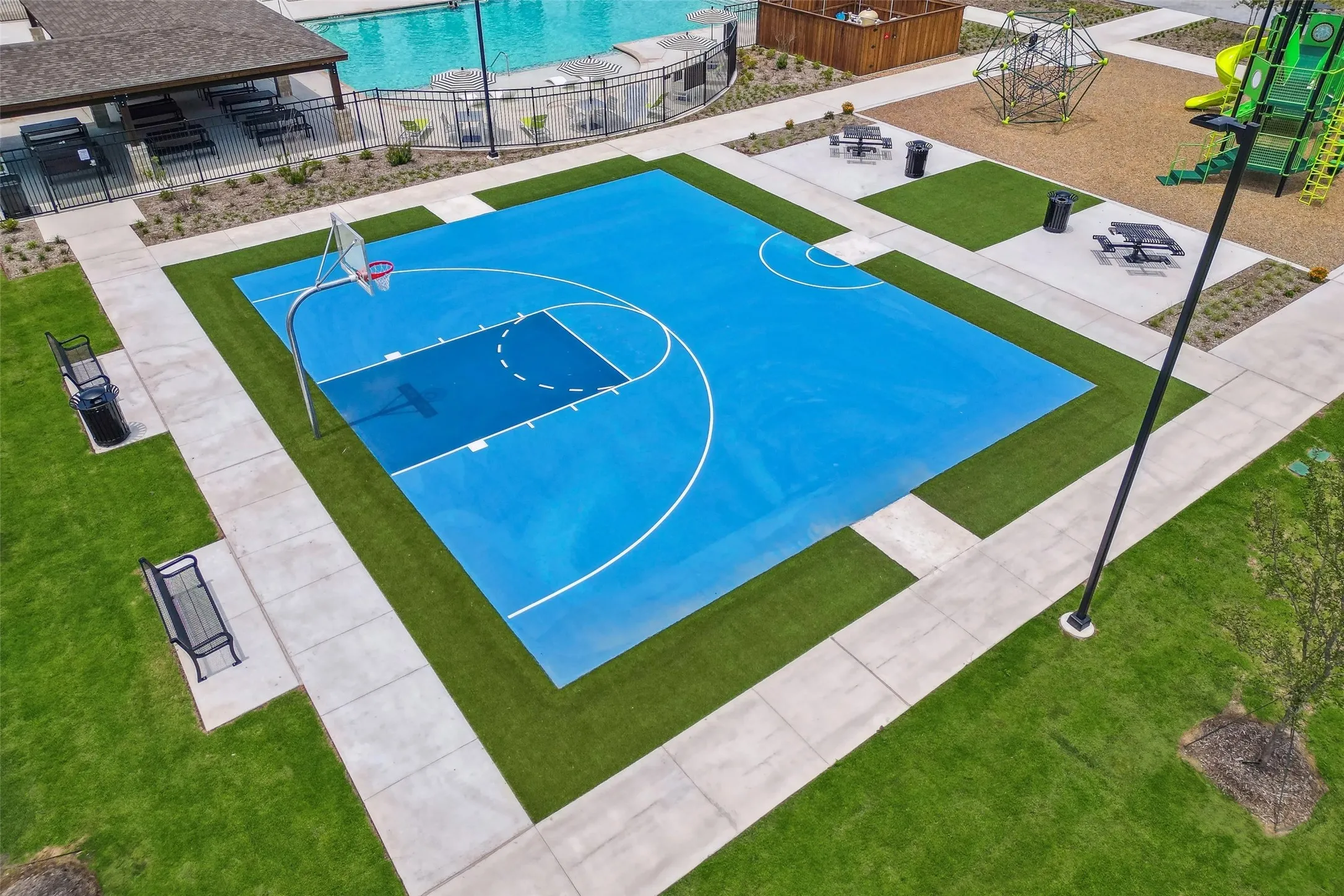 Outdoor basketball court with a vibrant blue surface, surrounded by pathways, artificial turf, and natural grass