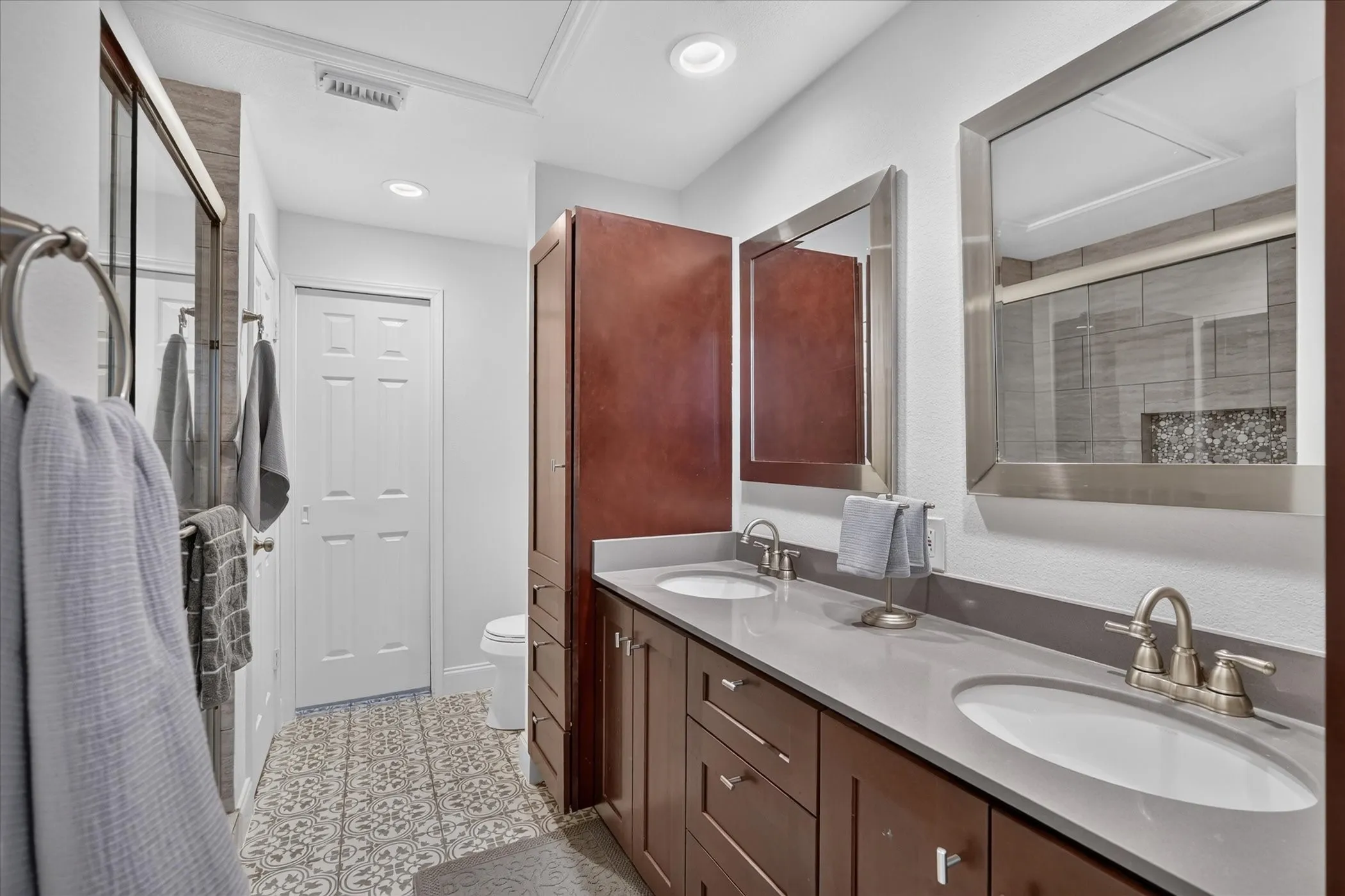 Dual sinks set in a gray countertop, dark wood-finish cabinetry, patterned floor tiling, and rectangular framed mirrors
