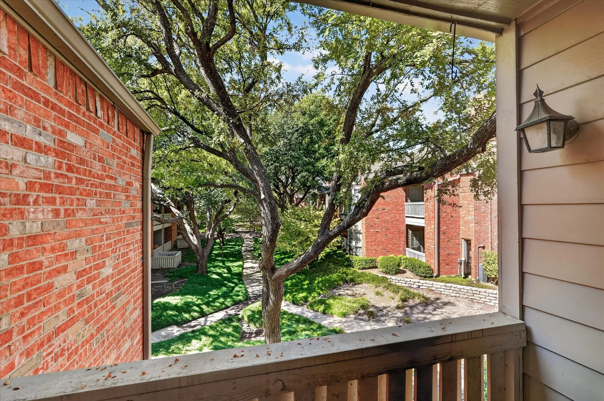 Balcony with wood railing overlooking a lush courtyard, featuring mature trees and brick building exteriors