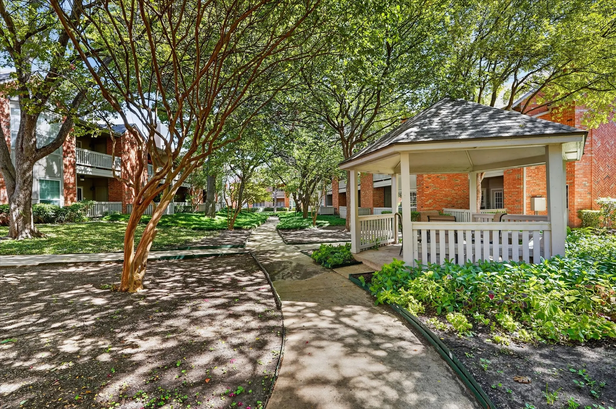 Manicured common grounds featuring mature trees, paved walking paths, a dedicated gazebo, and brick-exterior buildings