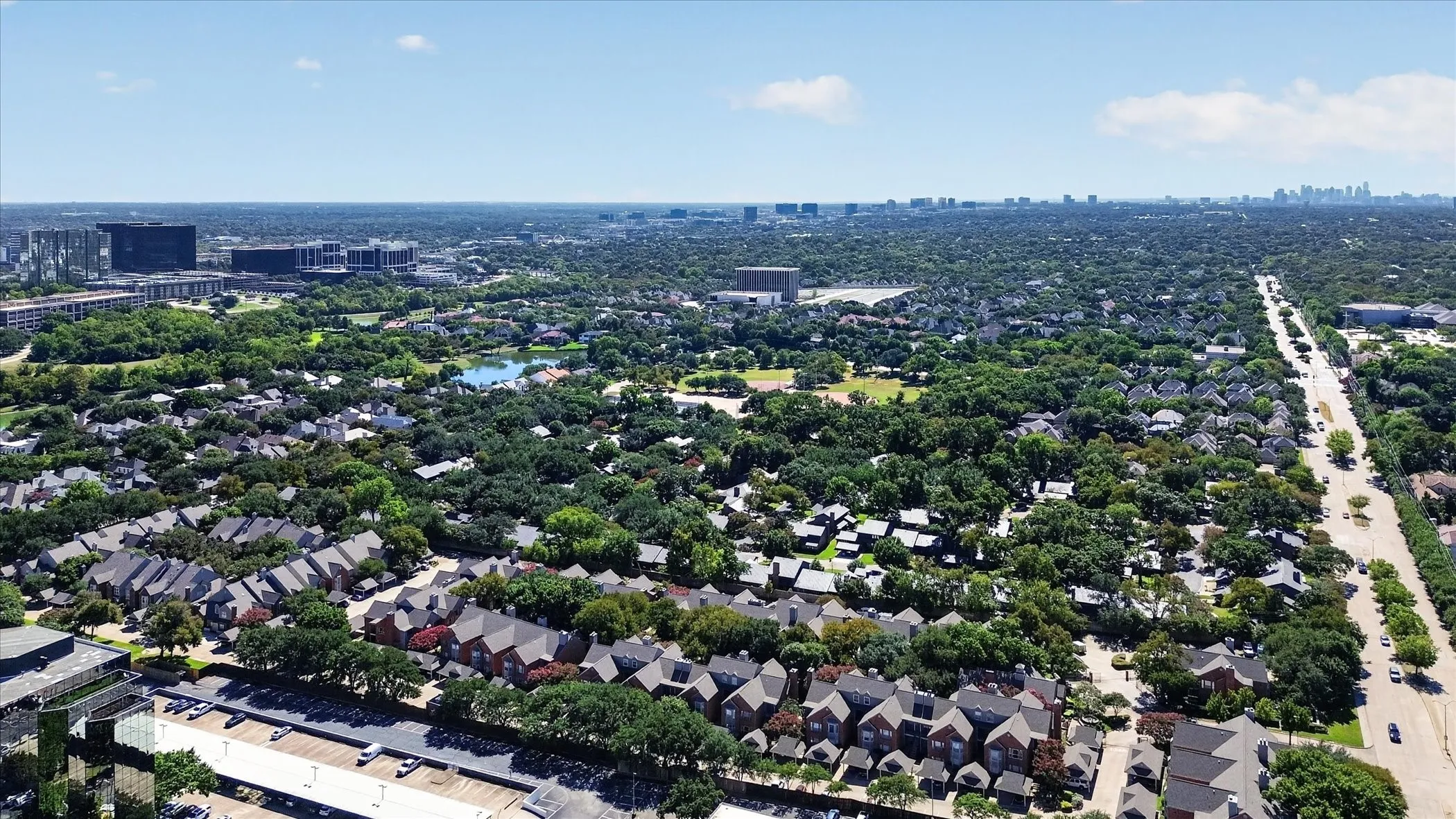 Expansive urban landscape featuring mature tree canopy, residential developments, a public roadway, a body of water, and commercial buildings