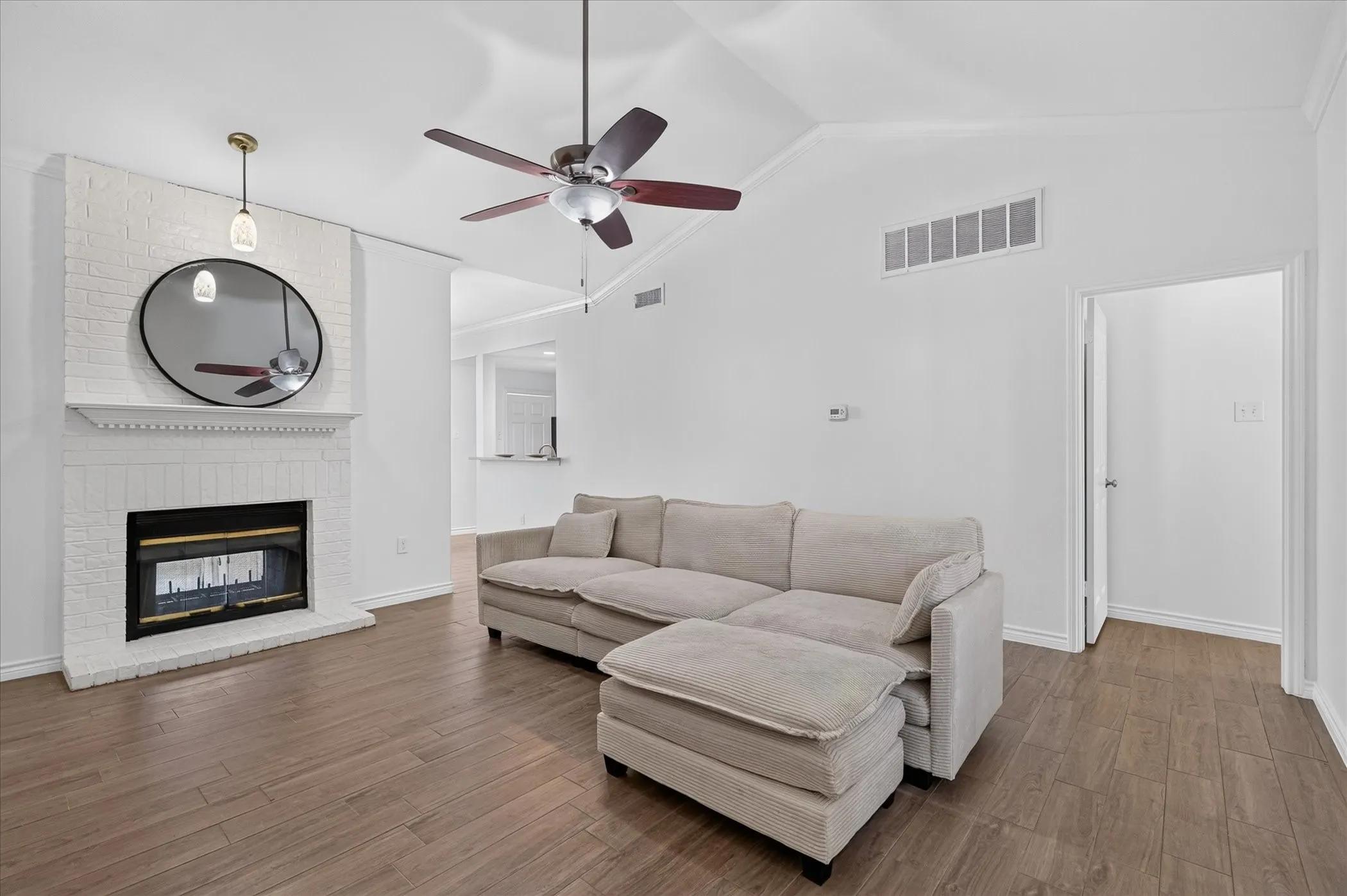 Vaulted ceiling with a ceiling fan, wood-finish flooring, white brick fireplace with a decorative mantel, and a doorway