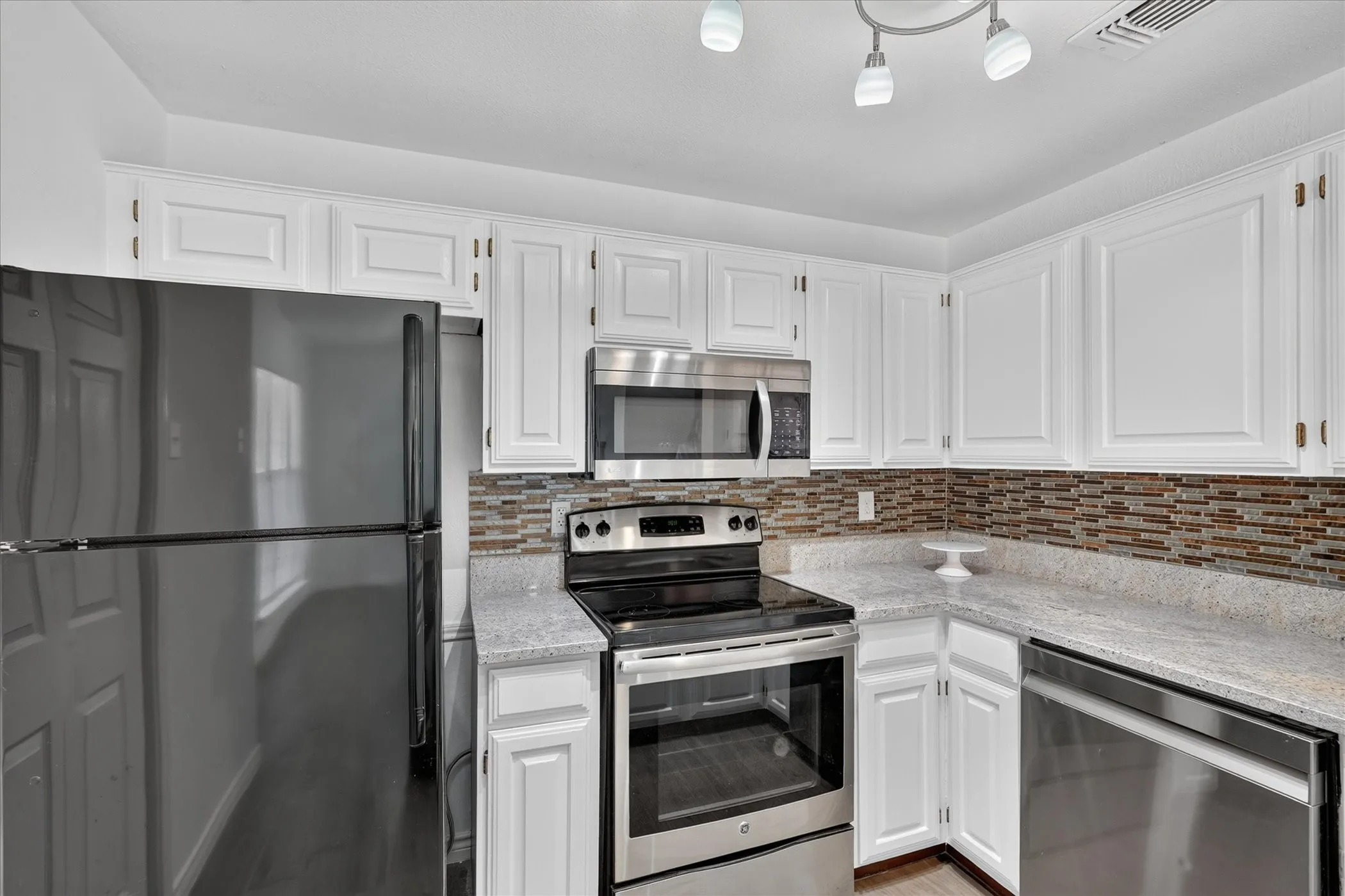 White cabinetry with panel detailing, mosaic tile backsplash, light-toned countertops, stainless steel appliances, and track lighting