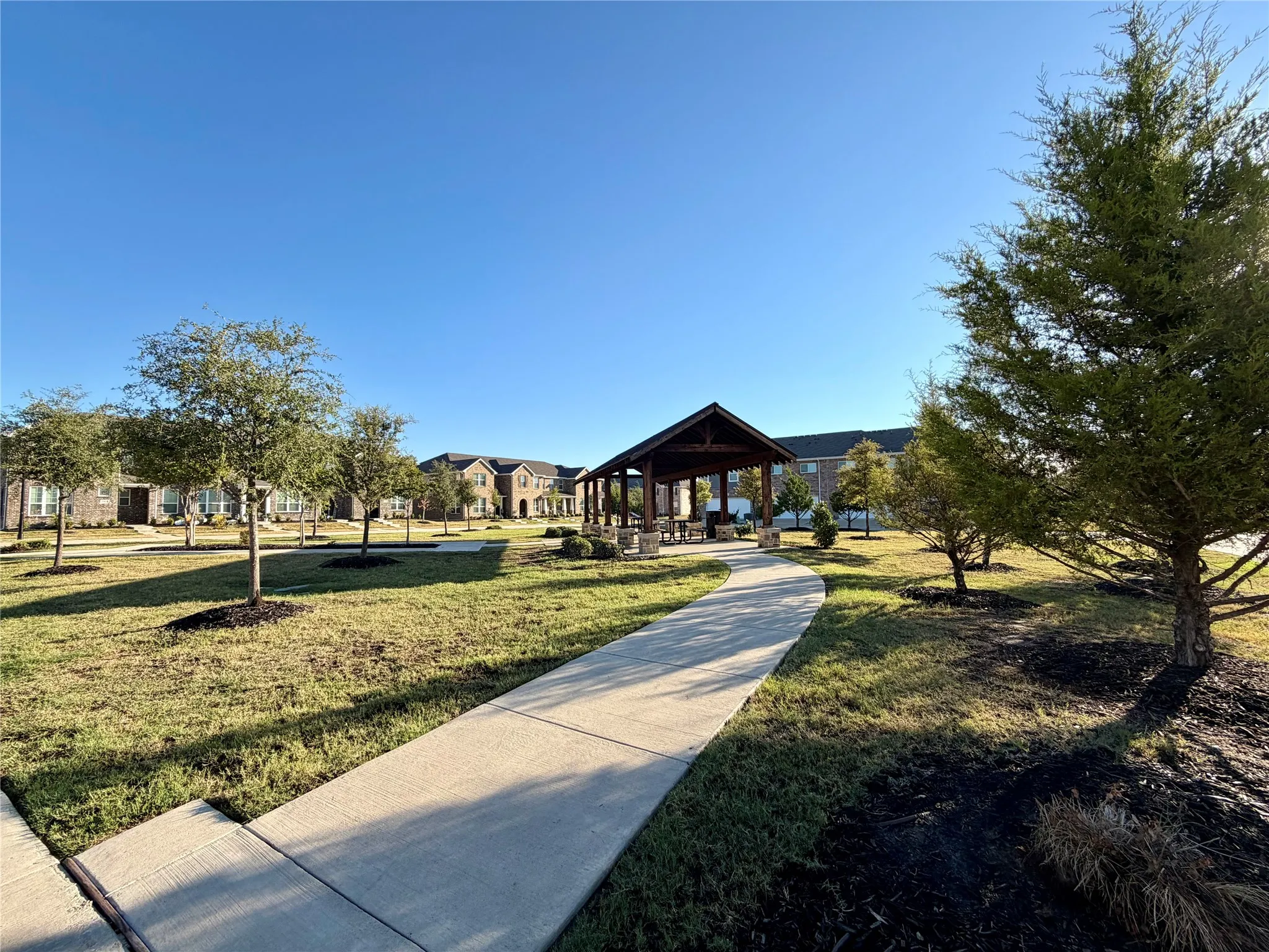 Community green space featuring a concrete pathway, a covered pavilion with stone pillars, and multiple mature trees