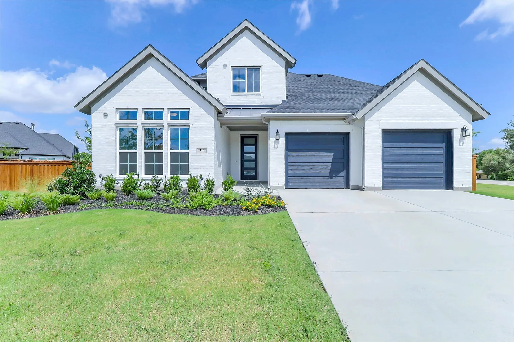 White brick exterior with a dark gray shingle roof, featuring a two-car garage with dark blue doors, a prominent front gable, and a concrete driveway