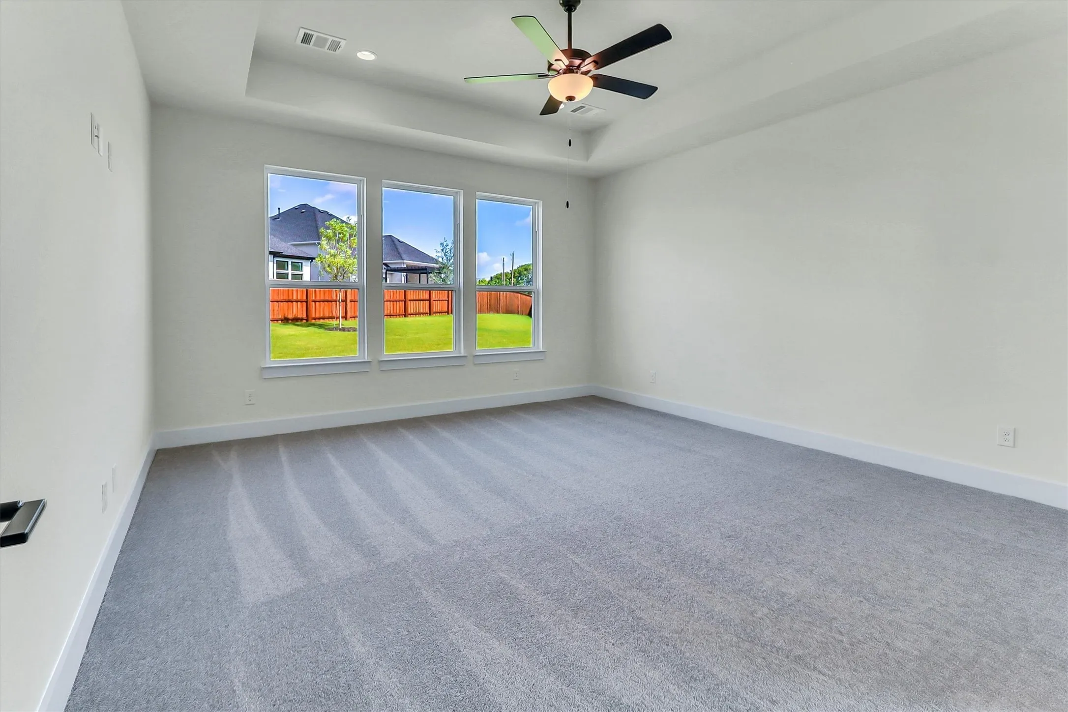 Spacious room featuring light gray carpeting, tray ceiling, ceiling fan with integrated lighting, and three large windows