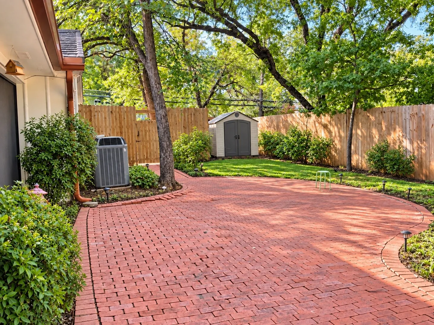 Expansive red brick paver patio with a curved edge, surrounded by mature landscaping and a wood fence