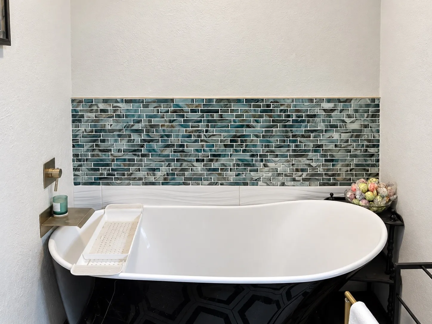 Freestanding soaking tub with a black hexagonal tiled exterior, complemented by a mosaic tile accent wall and a wall-mounted brass faucet
