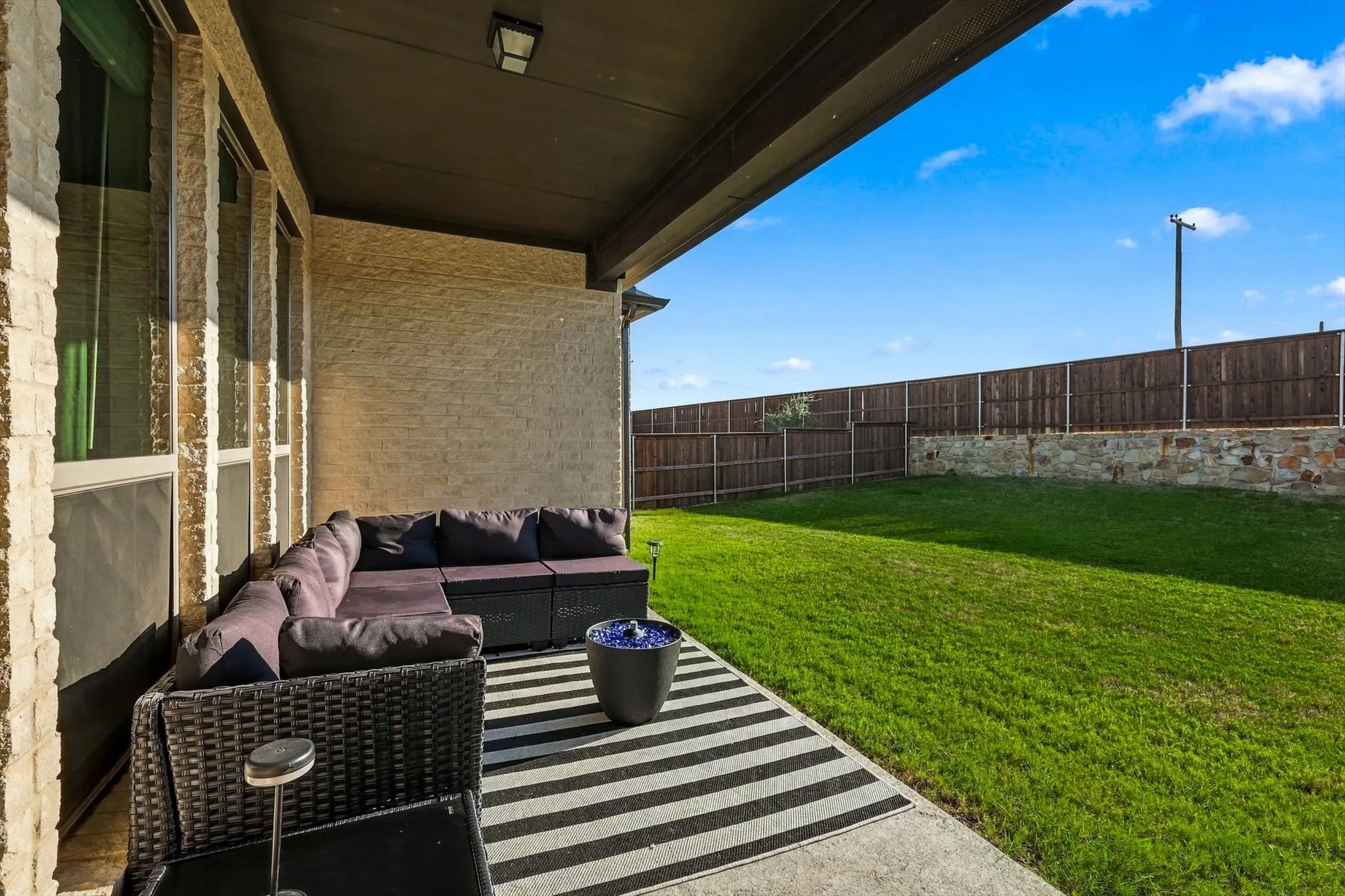 Covered patio with concrete flooring, stone veneer exterior, a spacious lawn area, and a wood privacy fence on a stone base