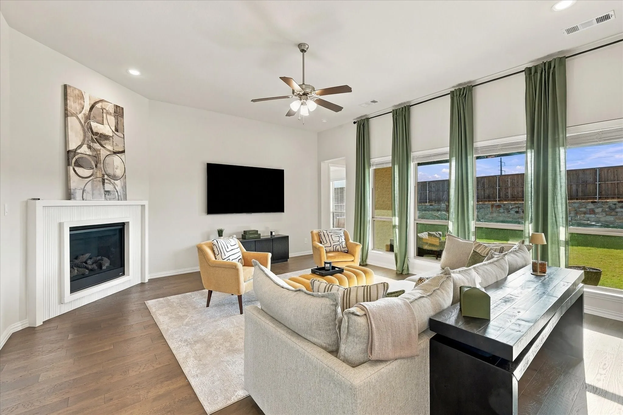 Spacious living area featuring wood-finish flooring, a white fireplace, a ceiling fan, and large windows with roller shades