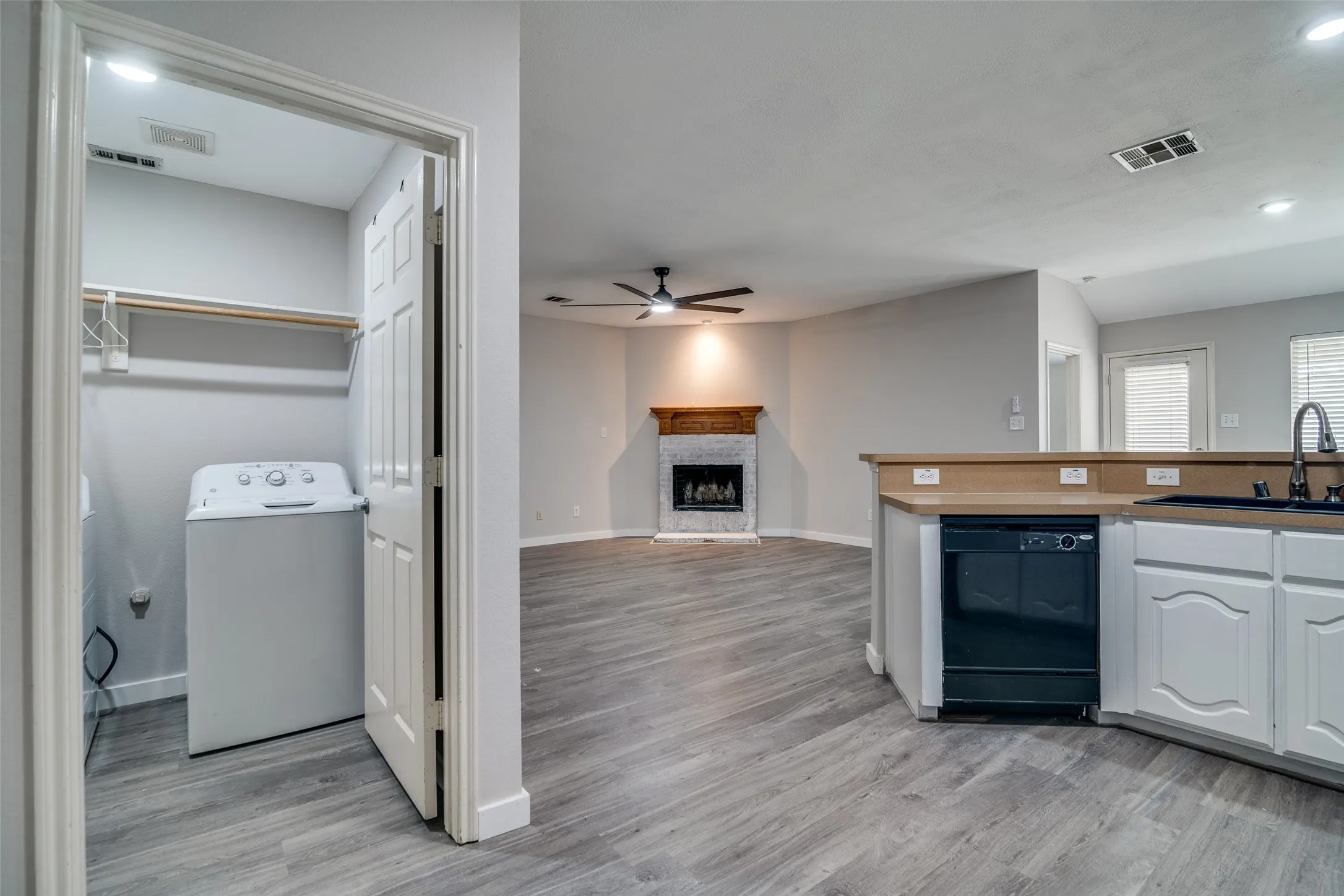 Laundry closet with built-in shelving, wood-finish flooring, corner fireplace with wood mantel, kitchen peninsula with integrated dishwasher, and ceiling fan