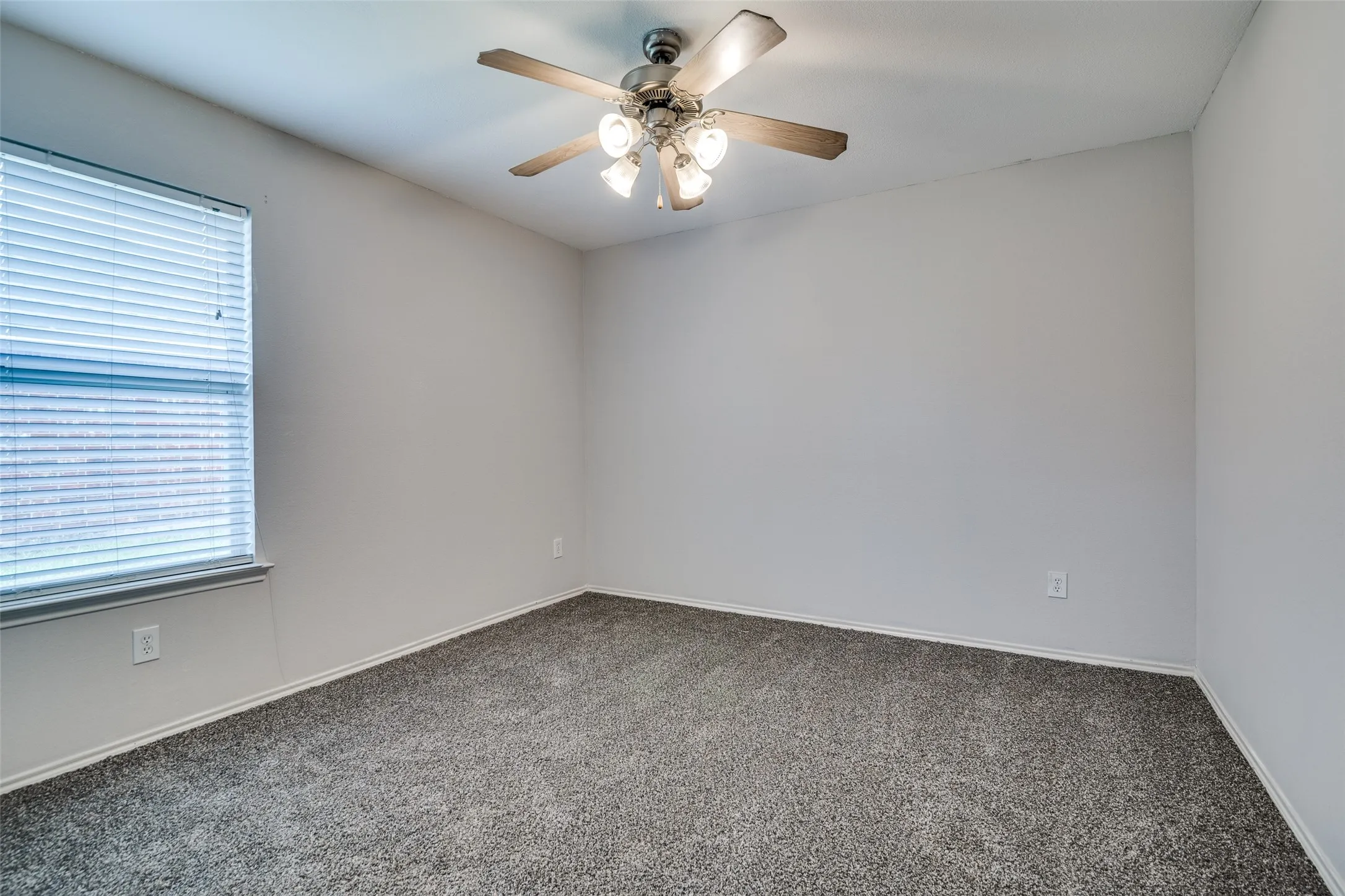 Neutral wall color, grey carpet flooring, window with blinds, and a ceiling fan with integrated lighting