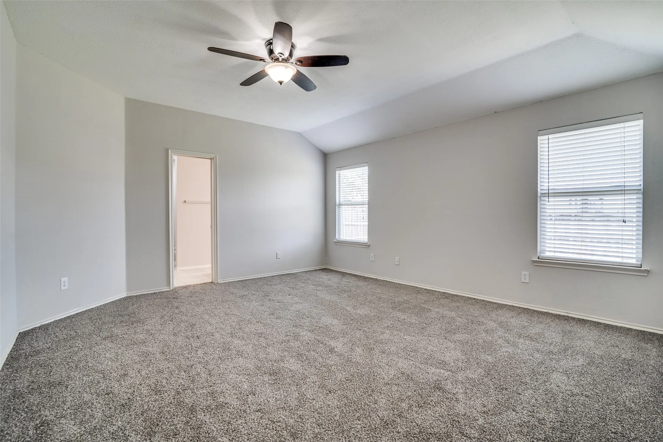Vaulted ceiling, two windows with blinds, ceiling fan with integrated lighting, neutral wall paint, and gray carpet flooring