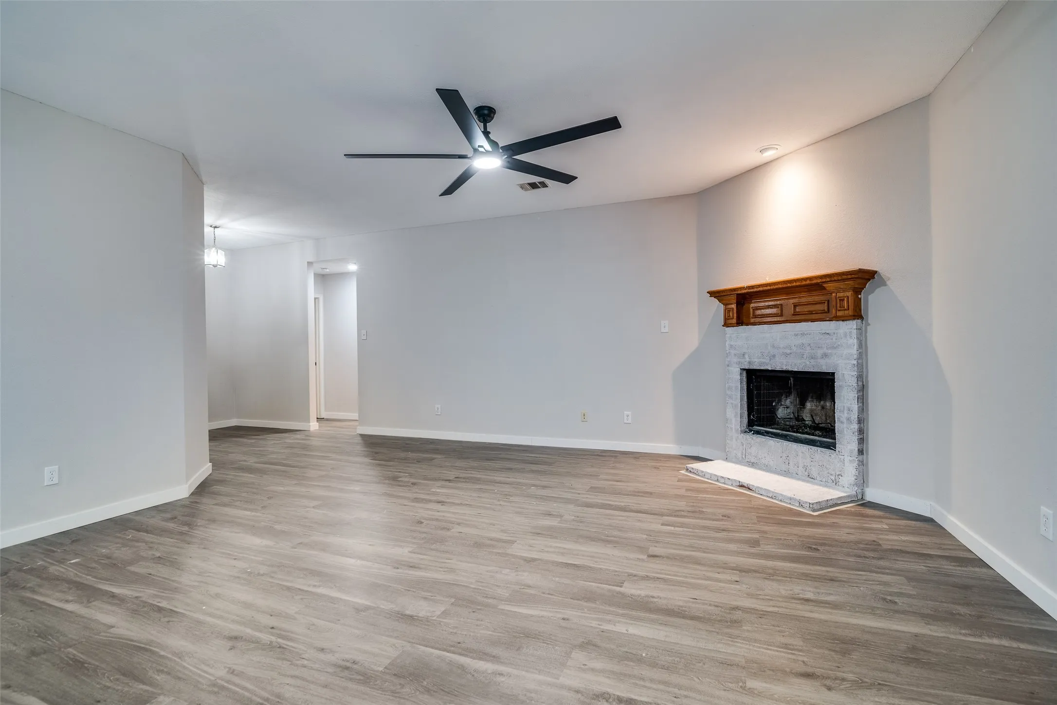 Spacious living area featuring wood-finish flooring, a contemporary ceiling fan, and a brick fireplace with a wood mantle and hearth