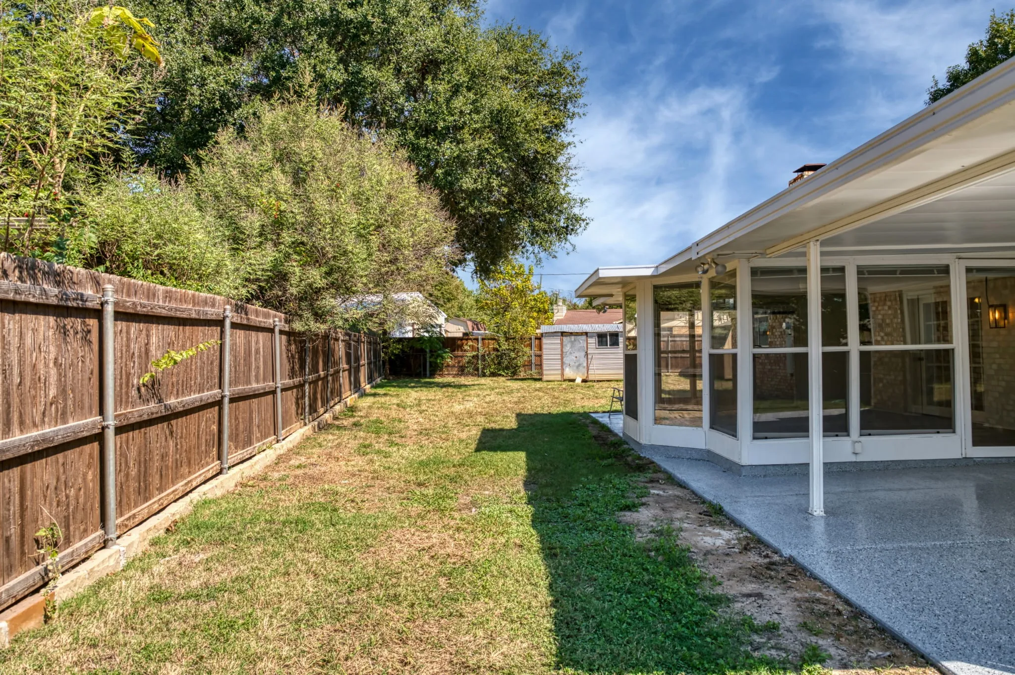 Expansive backyard featuring a wood privacy fence, a covered patio with a textured concrete surface, and a screened sunroom