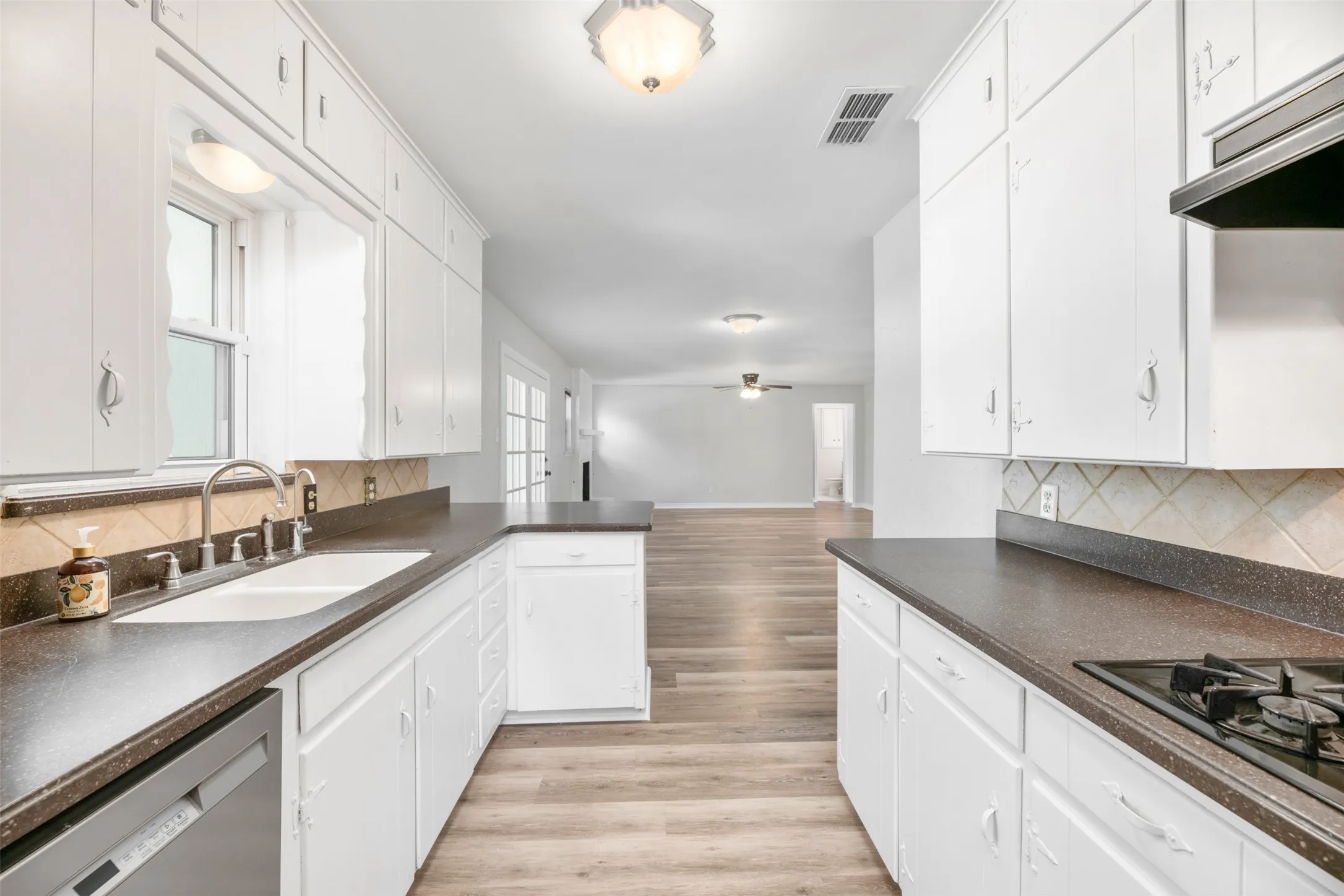 Galley kitchen featuring white cabinetry, dark countertops, a double-basin sink, stainless steel dishwasher, and tile backsplash