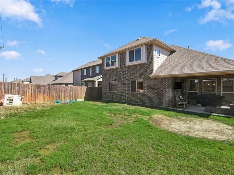 Expansive backyard featuring a manicured lawn with mature landscaping, wooden privacy fencing, and a brick exterior with multiple gabled rooflines