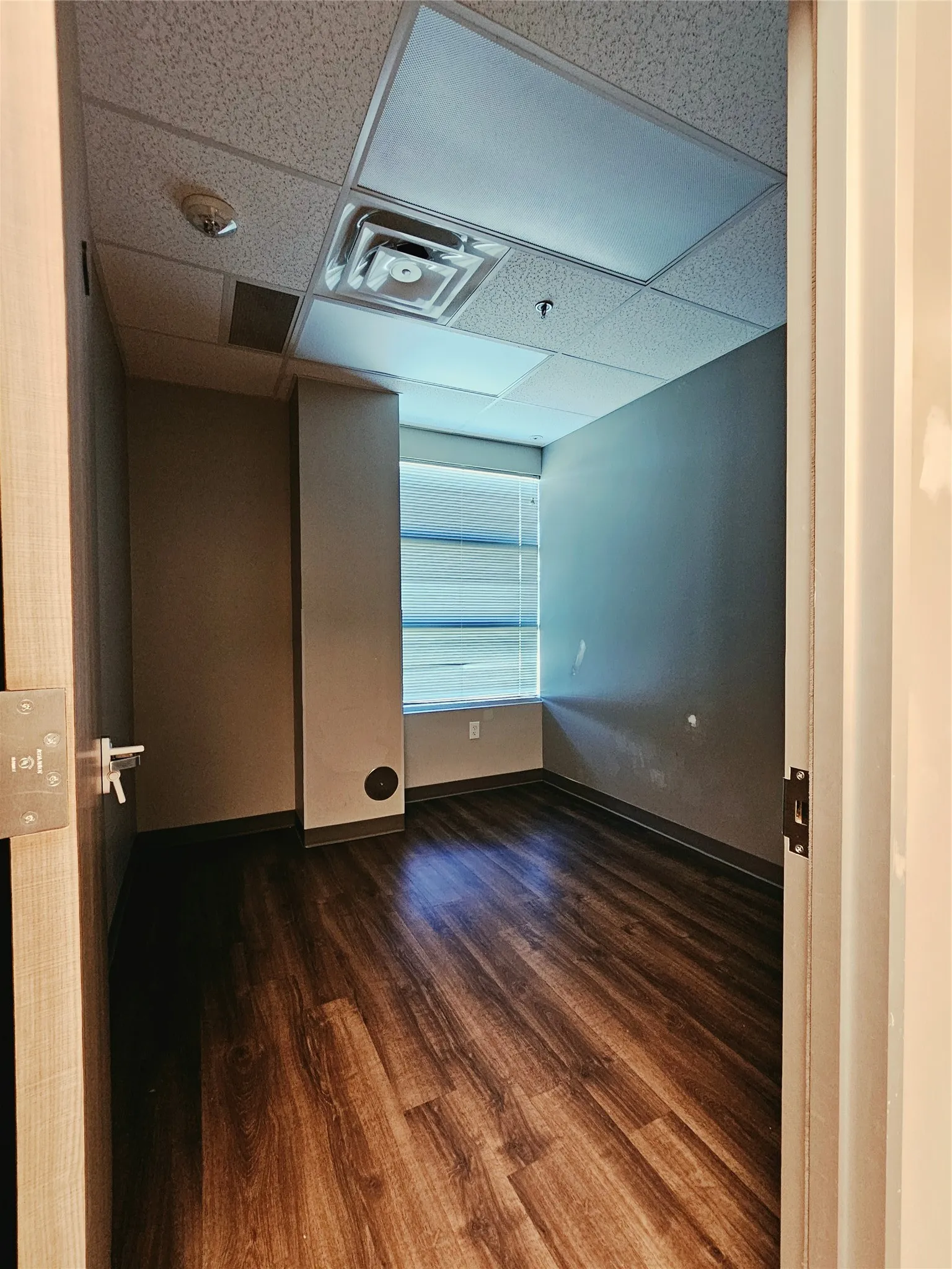 Wood-finish flooring, drop ceiling with integrated lighting, and a window with horizontal blinds