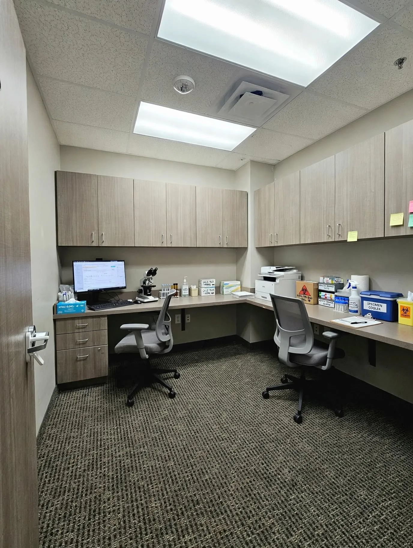 Corner office workspace featuring L-shaped countertop, under-cabinet storage, wood-finish cabinetry, and overhead fluorescent lighting