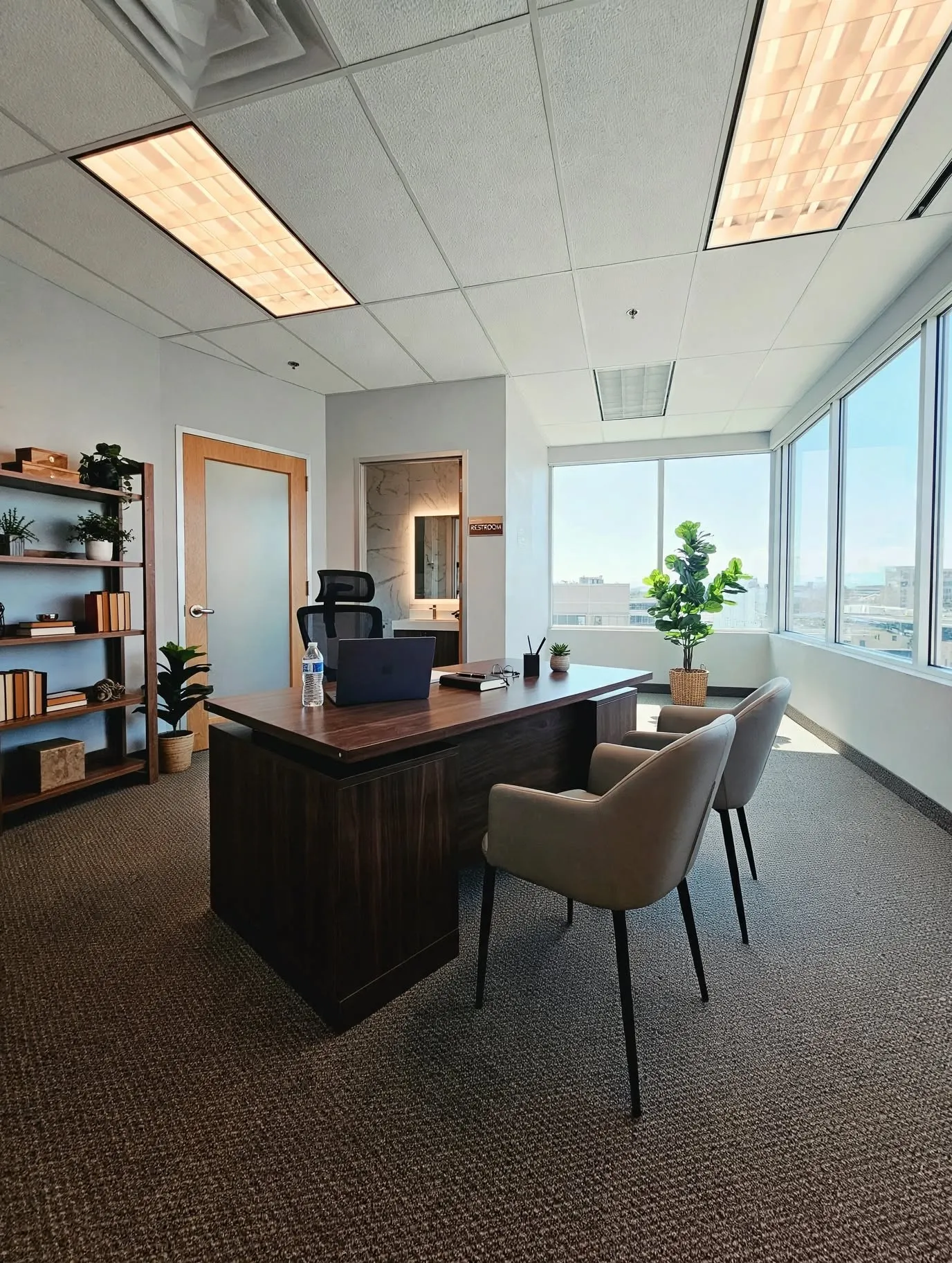 Wood-finish executive desk with integrated storage, full-height windows, recessed ceiling lighting, and carpeted flooring