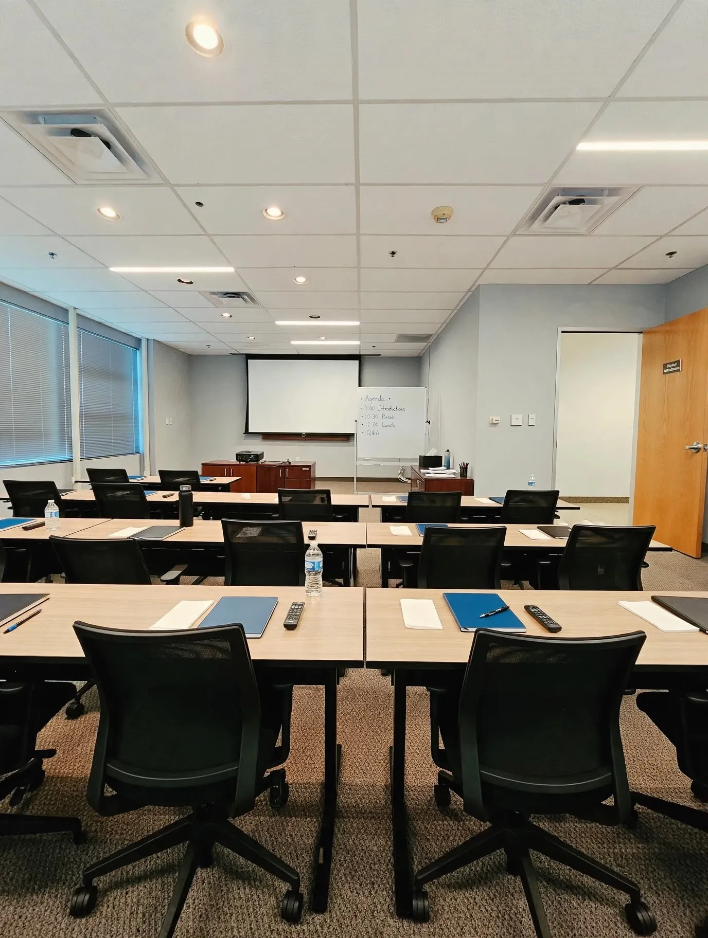 Modern training or conference room featuring multiple workstations with ergonomic black task chairs, light wood-finish tables, and abundant natural light from floor-to-ceiling windows