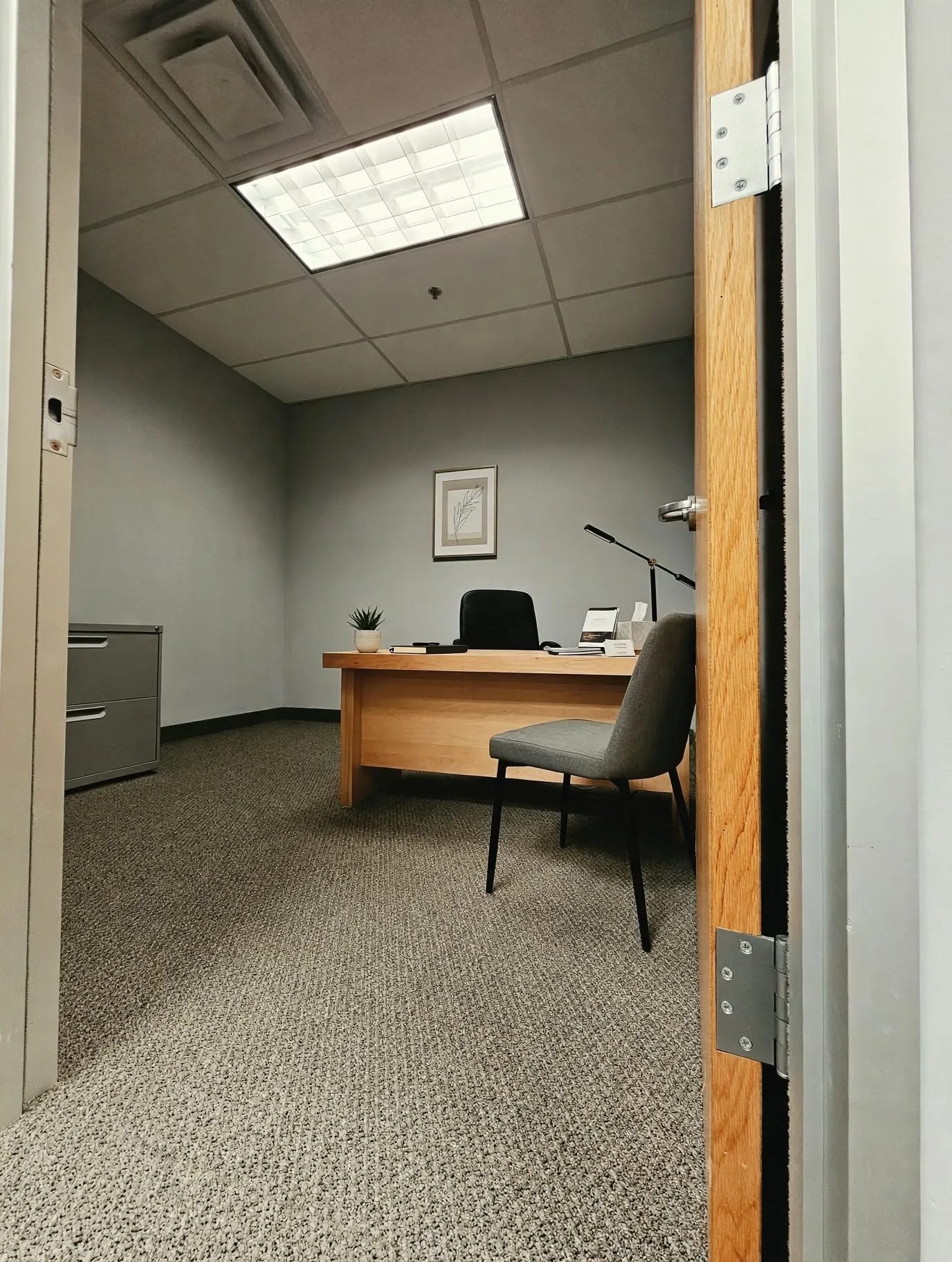 Textured carpet flooring, suspended ceiling with recessed lighting, light wood-finish door, and light gray wall paint