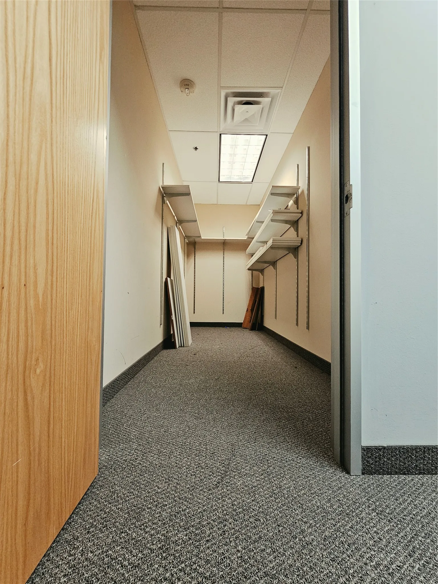 Recessed lighting, acoustic ceiling tiles, light wood-finish door, carpet flooring, wall-mounted shelving brackets
