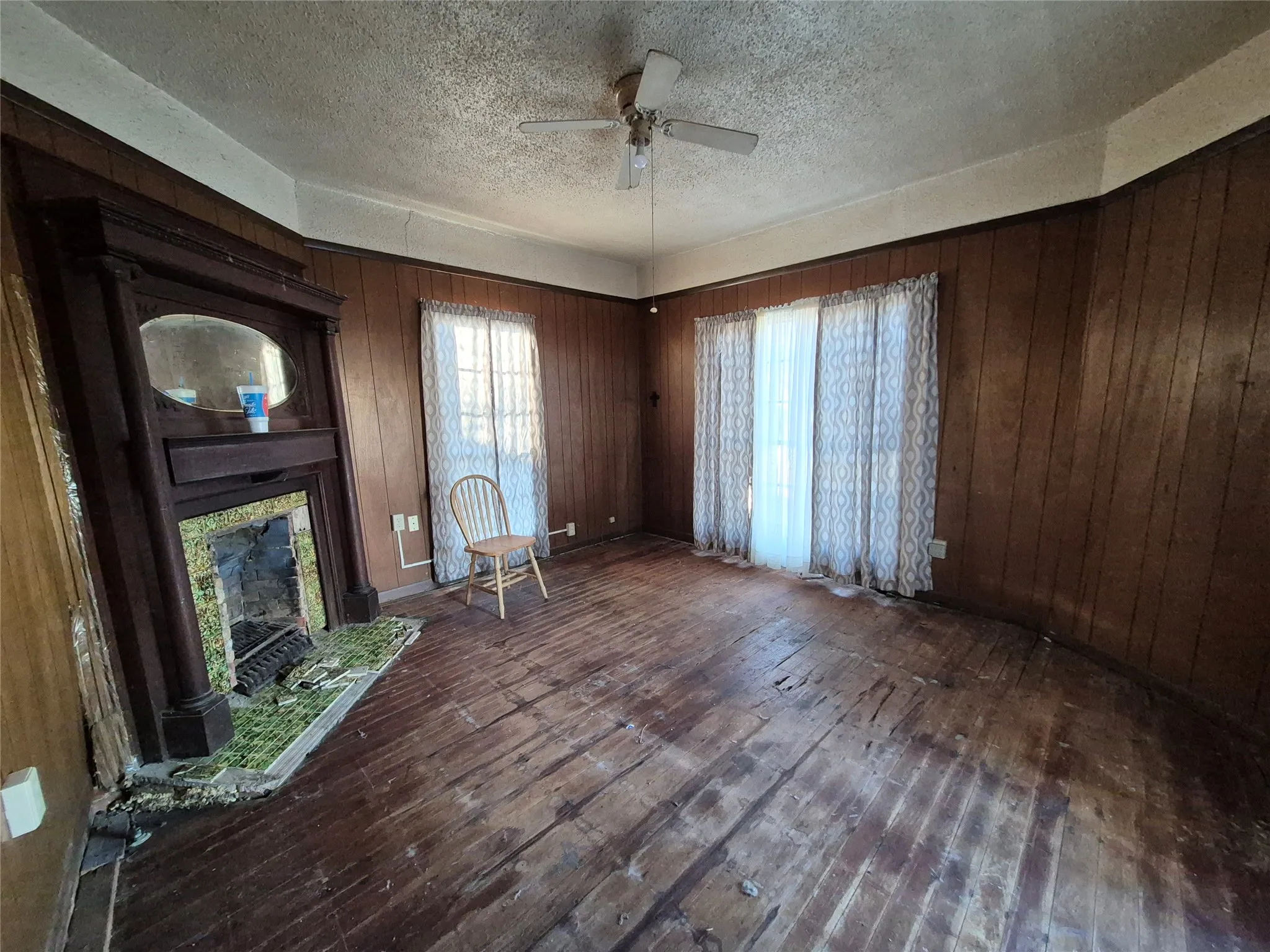 Wood paneling walls, wood-finish flooring, two window openings, ceiling fan, and a fireplace with a wood mantel