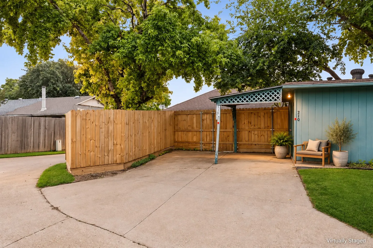 Expansive concrete driveway with a wooden privacy fence, mature trees, and a covered carport with lattice detailing