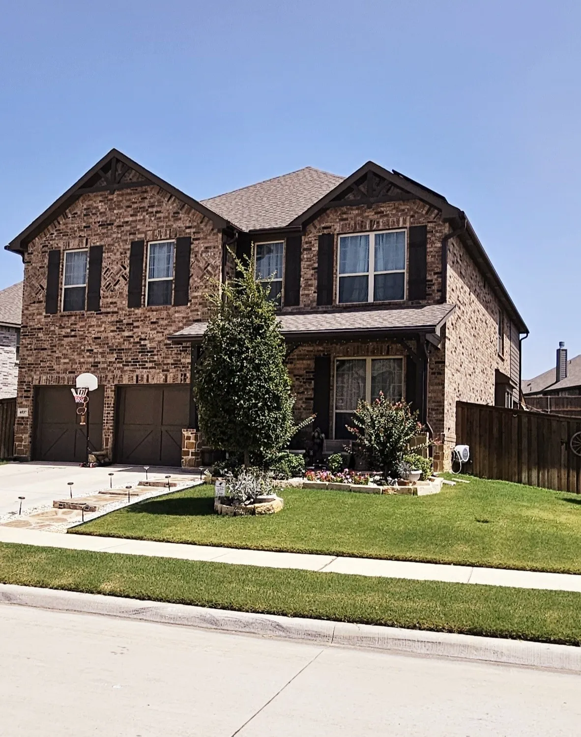 Brick exterior with dark shutters, dual garage doors, a sloped shingle roof, and a mature tree in the front yard