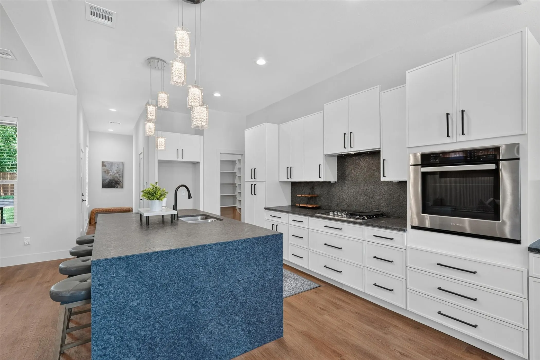 Kitchen featuring wood-finish flooring, white cabinetry with black hardware, dark stone countertops, a built-in wall oven, and a kitchen island with a sink and pendant lighting