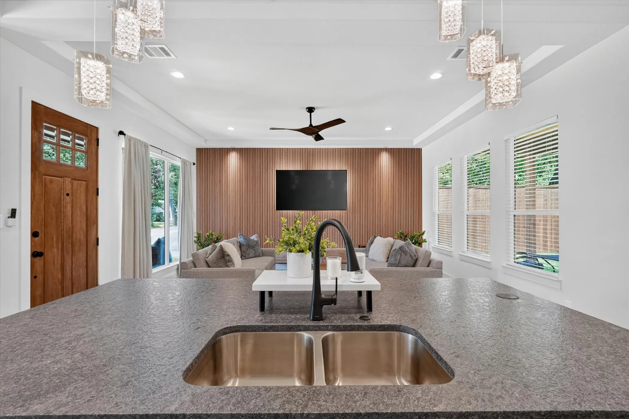 Dual basin stainless steel sink, dark textured countertop, wood-finish accent wall, ceiling fan, and multiple pendant light fixtures