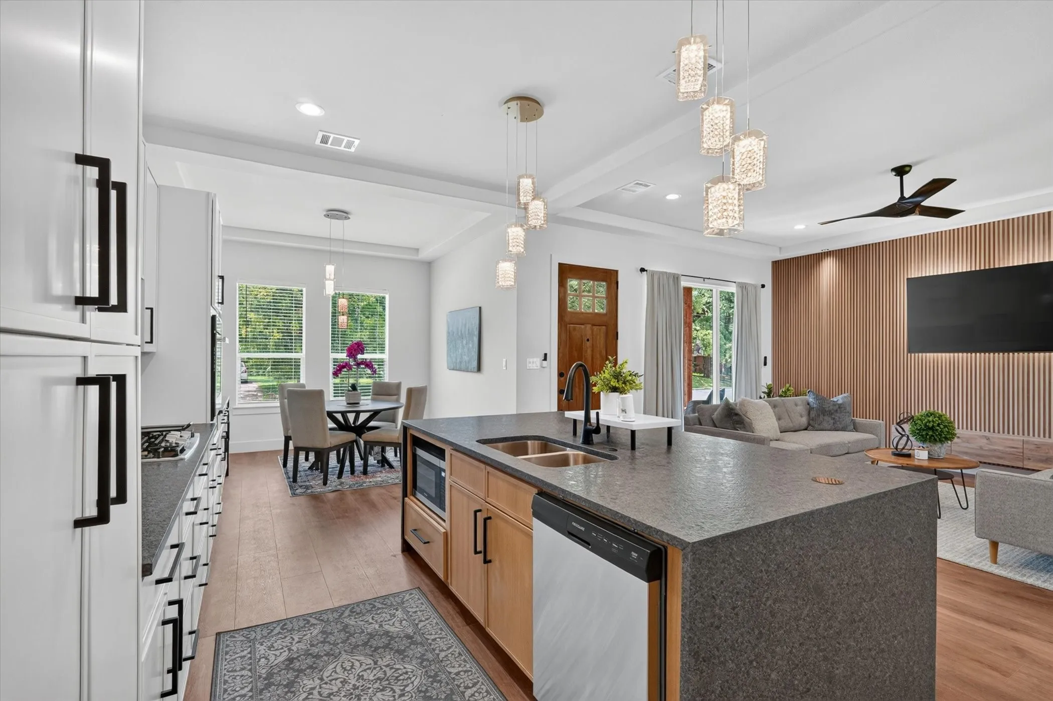 Spacious kitchen island with an integrated sink and dishwasher, white cabinetry with black hardware, wood-finish flooring, and a dining area with pendant lighting