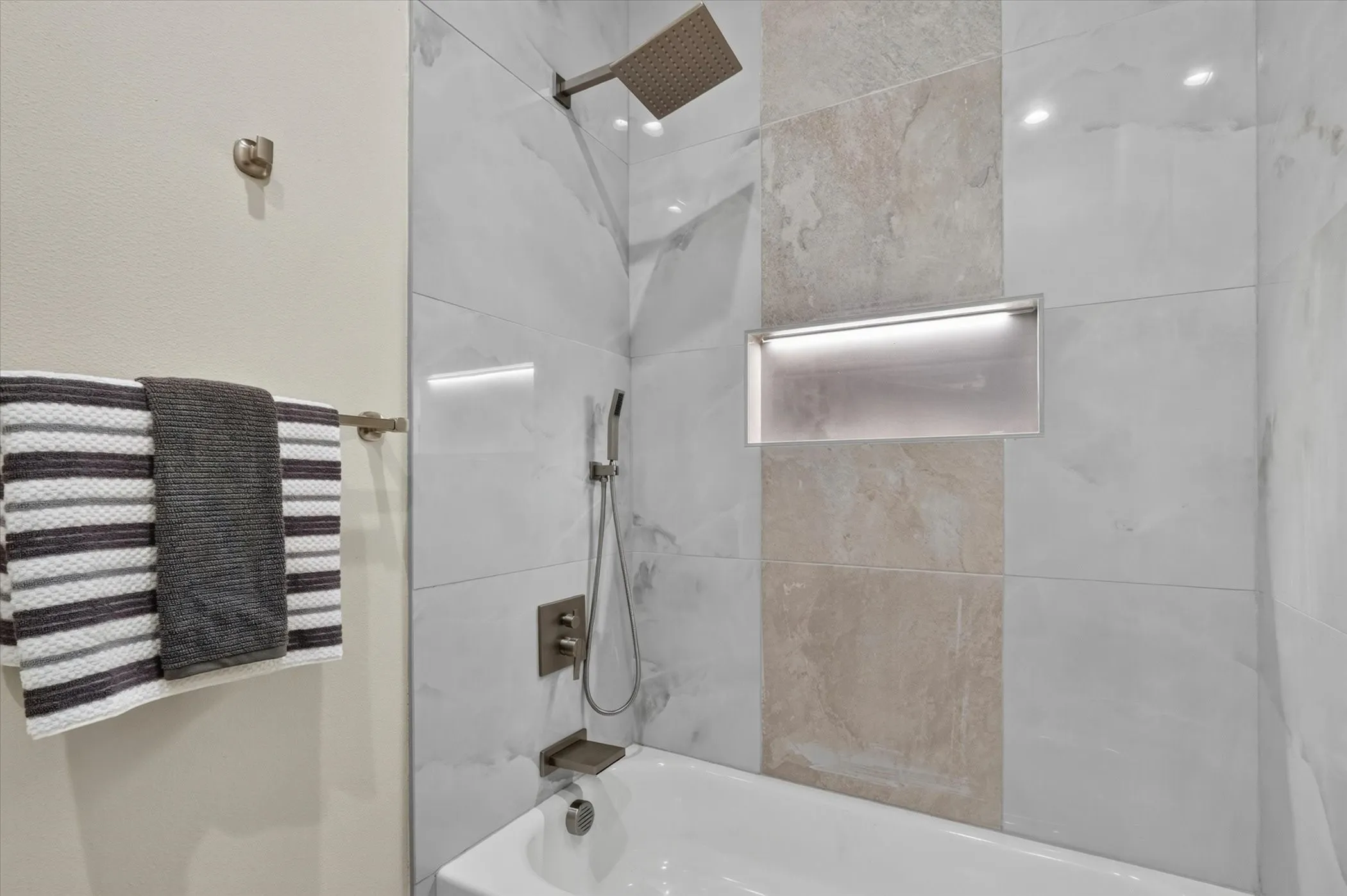 Bathtub with mixed tile surround, integrated shower niche with accent lighting, and a rain shower head with a handheld sprayer