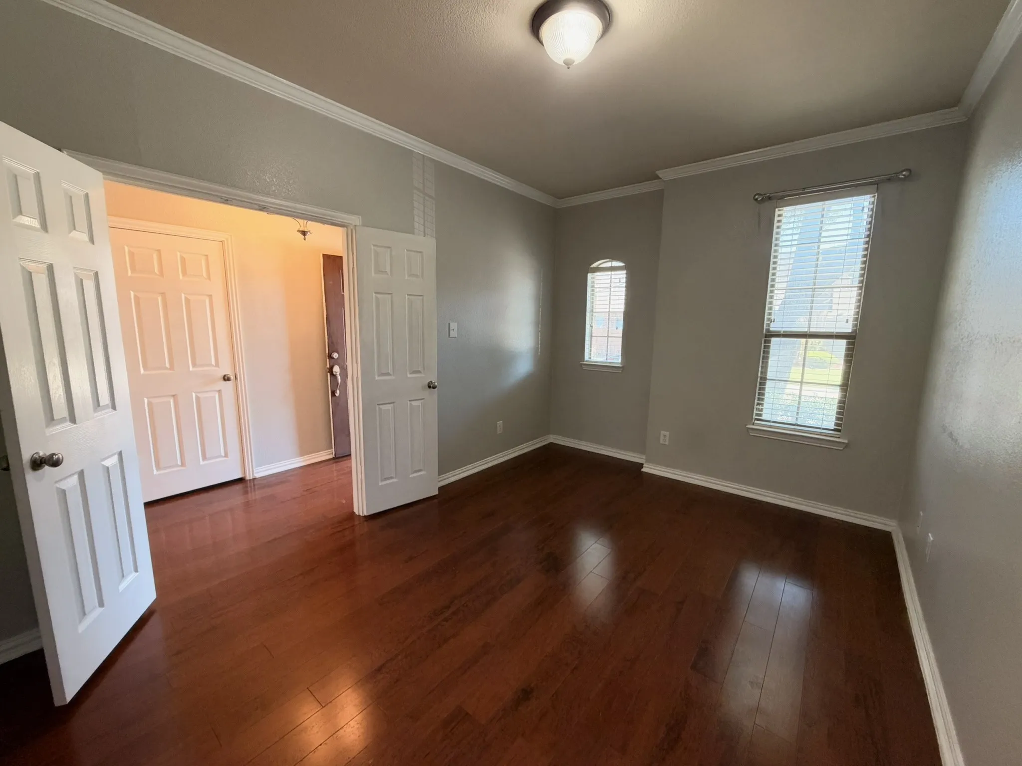 Rich wood-finish flooring, two large windows with blinds, a compact arched window, a ceiling-mounted light fixture, and crown molding