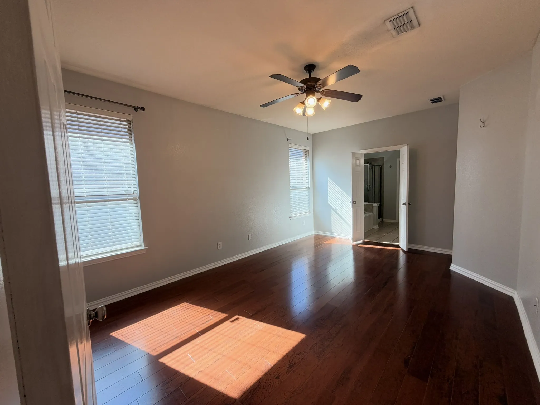 Dark wood-finish flooring, two window openings with blinds, ceiling fan with integrated lighting, and an open doorway revealing a bathtub