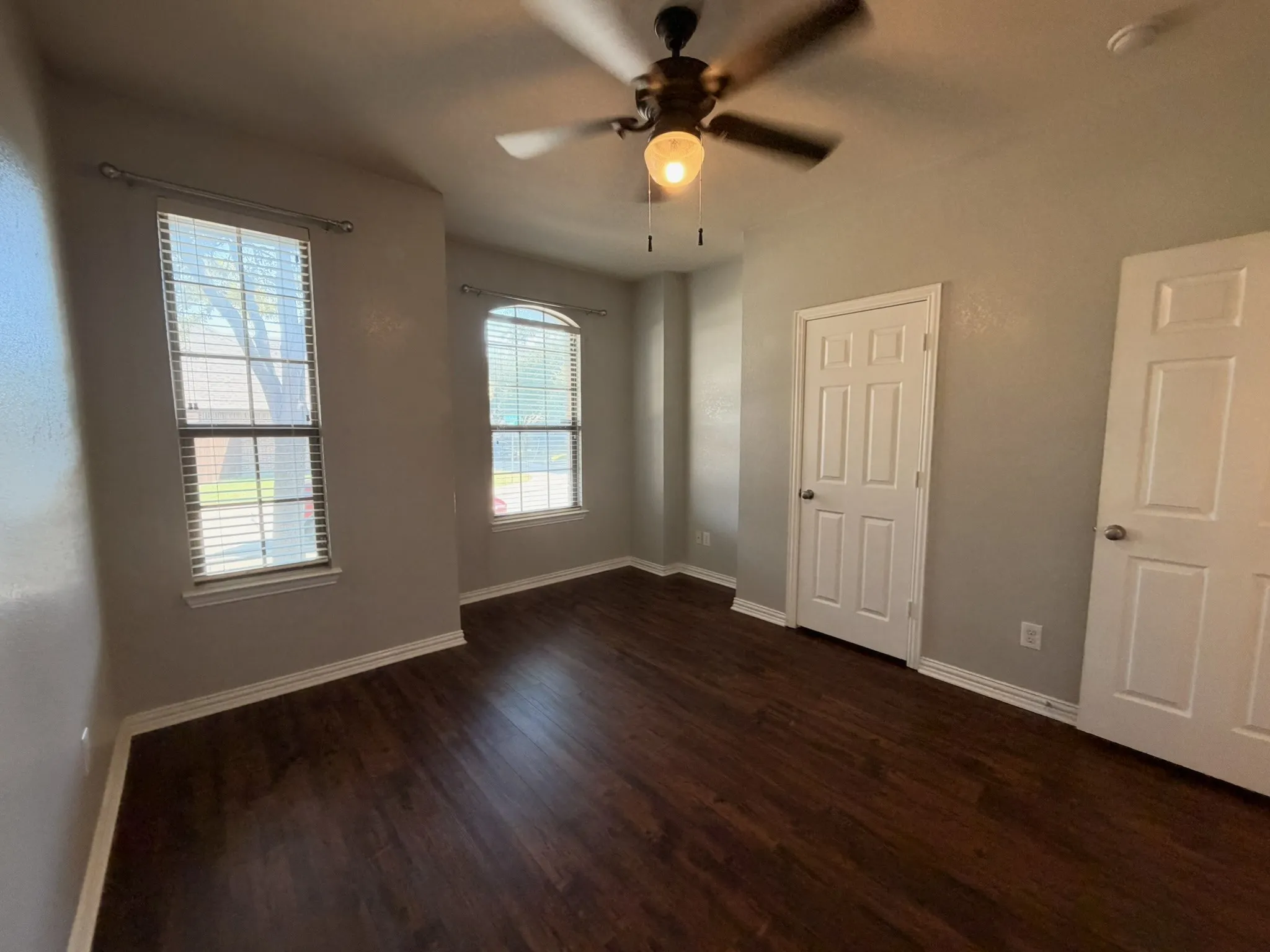 Wood-finish flooring throughout, two panel doors, two windows with blinds, and a ceiling fan with integrated lighting