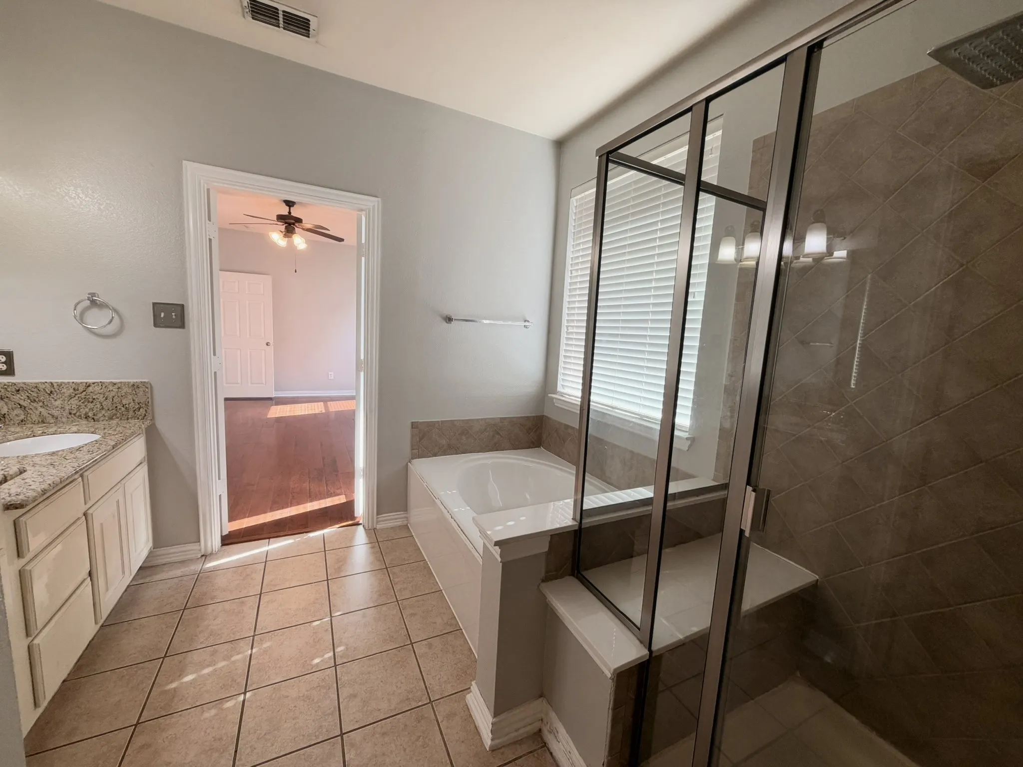 Tiled flooring, dual vanity with stone countertops, built-in soaking tub, separate glass-enclosed shower with tiled surround, and a window with blinds