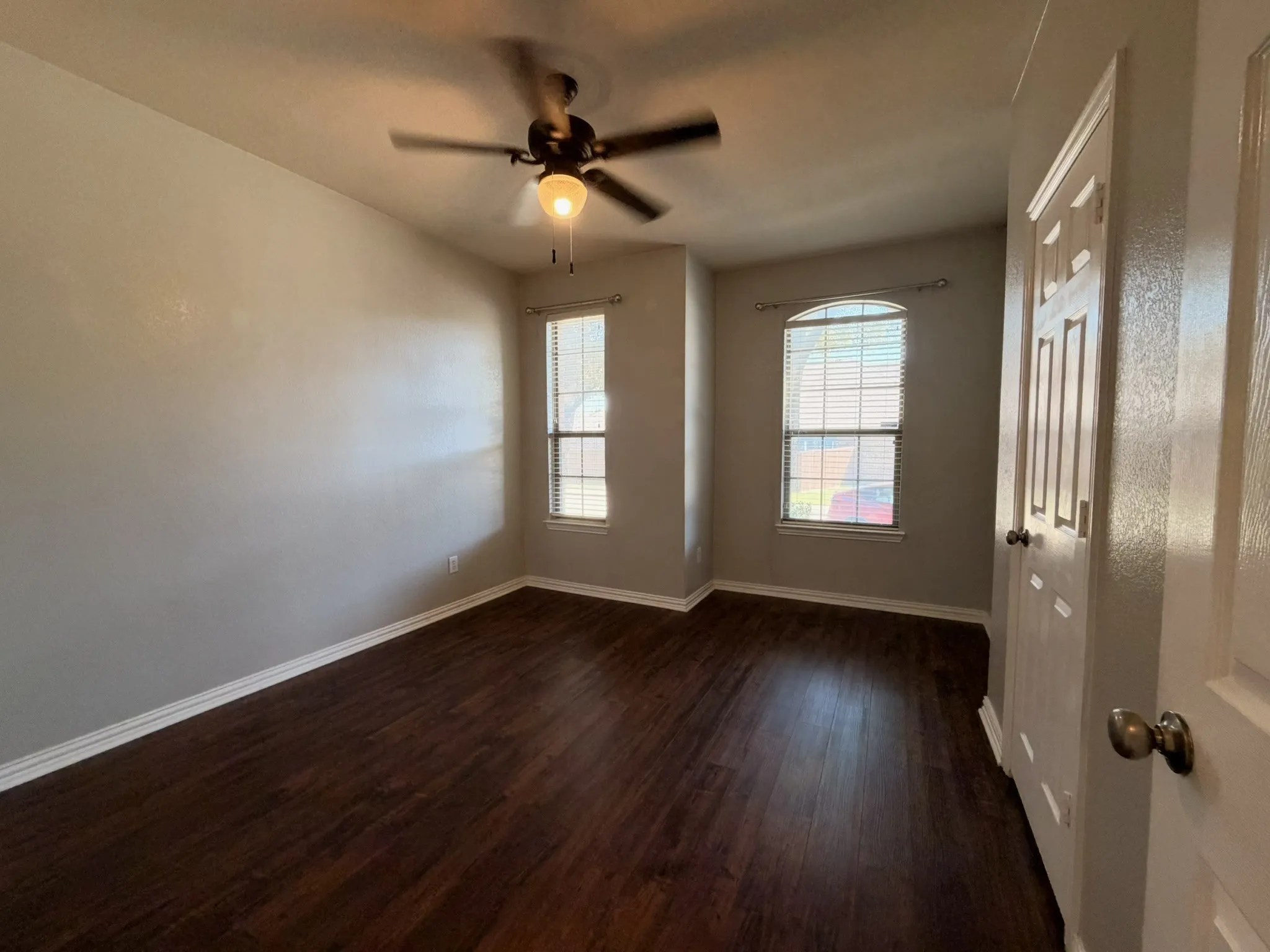 Rich wood-finish flooring, two large windows with blinds, and a ceiling fan with integrated lighting