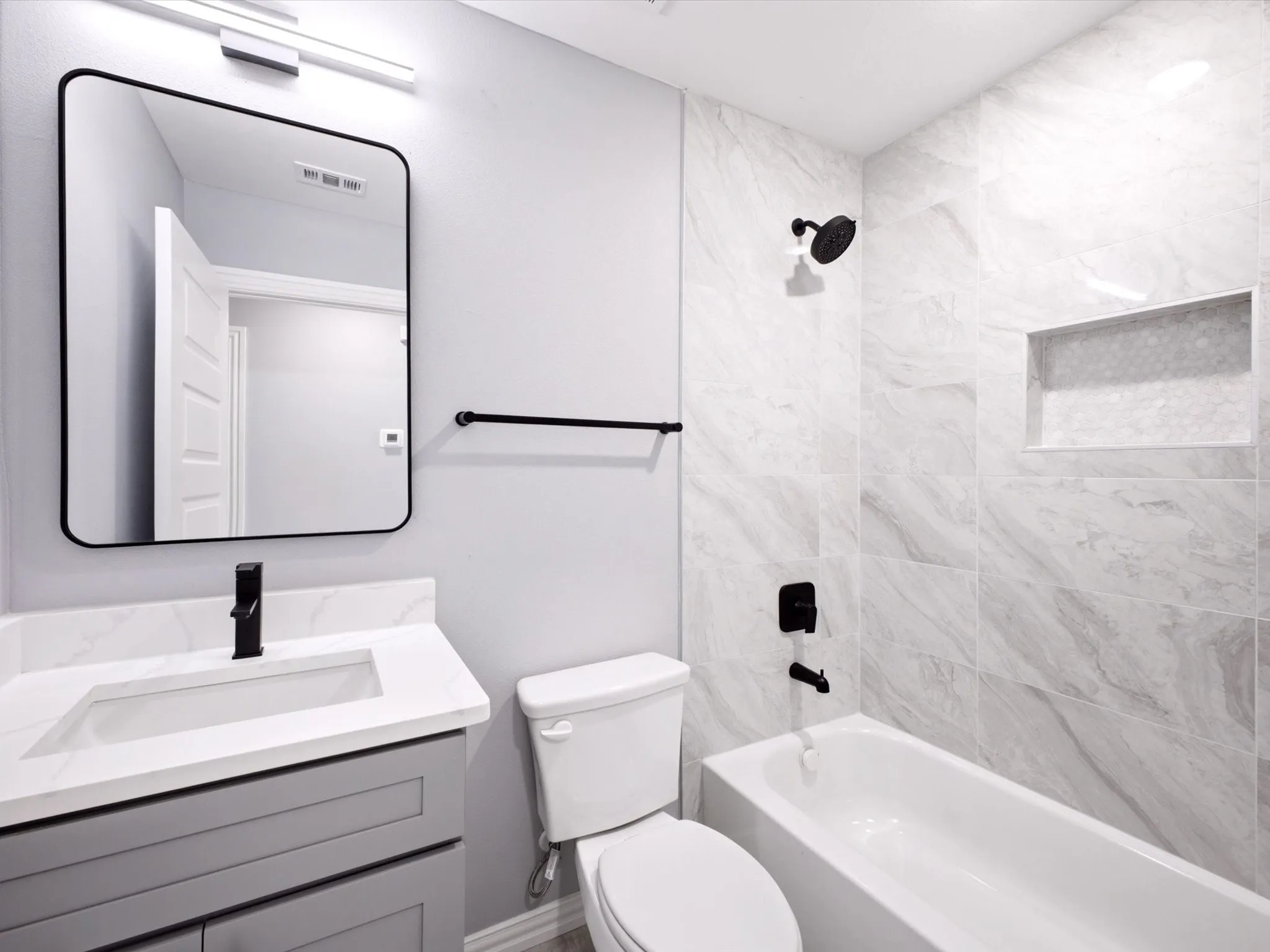White tiled tub and shower enclosure with a recessed shelf, black shower fixtures, a gray vanity with an integrated sink, and a black-framed mirror