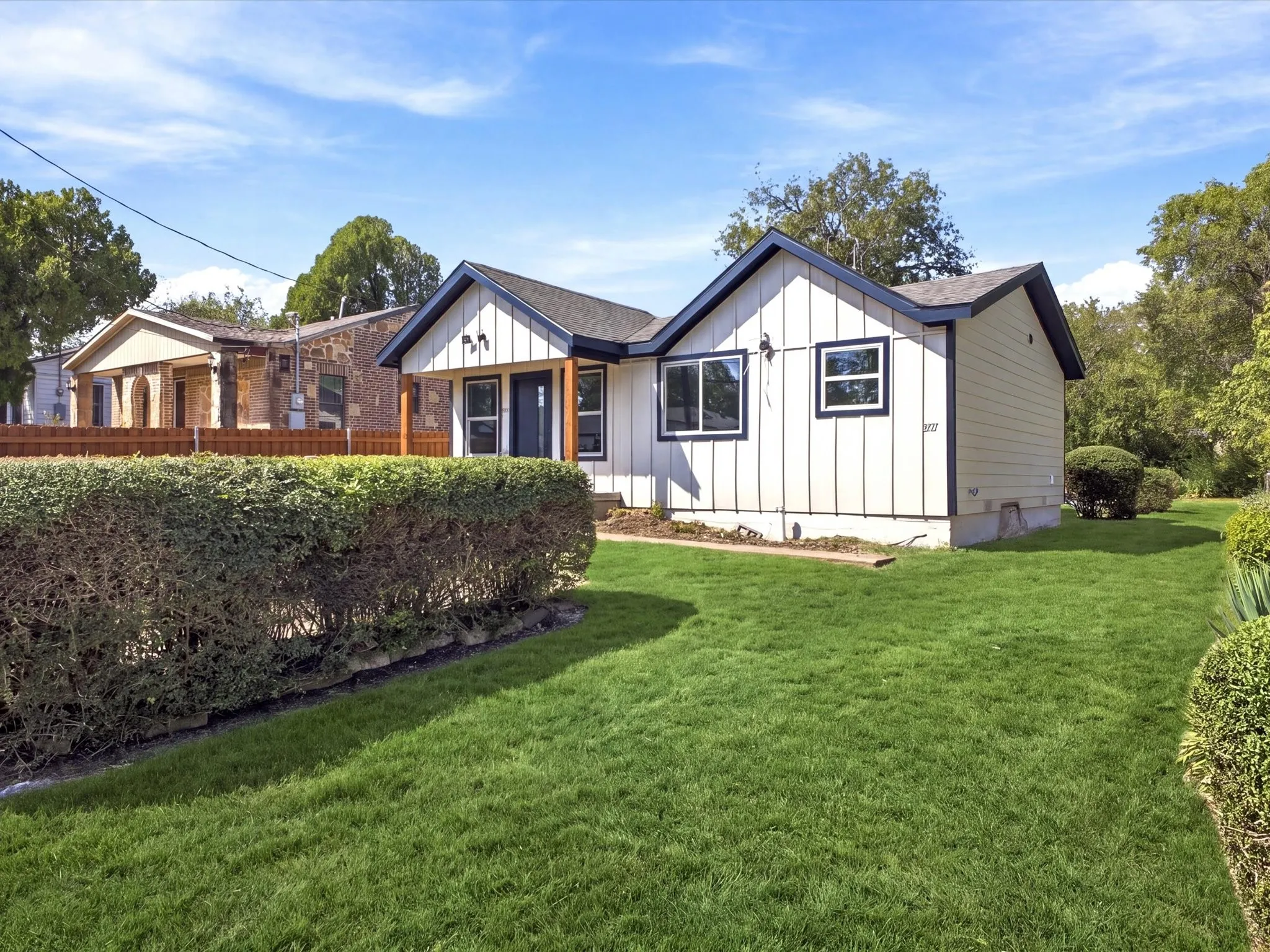 White exterior with dark trim accents, covered entry with wood posts, front yard with manicured lawn, and mature hedges
