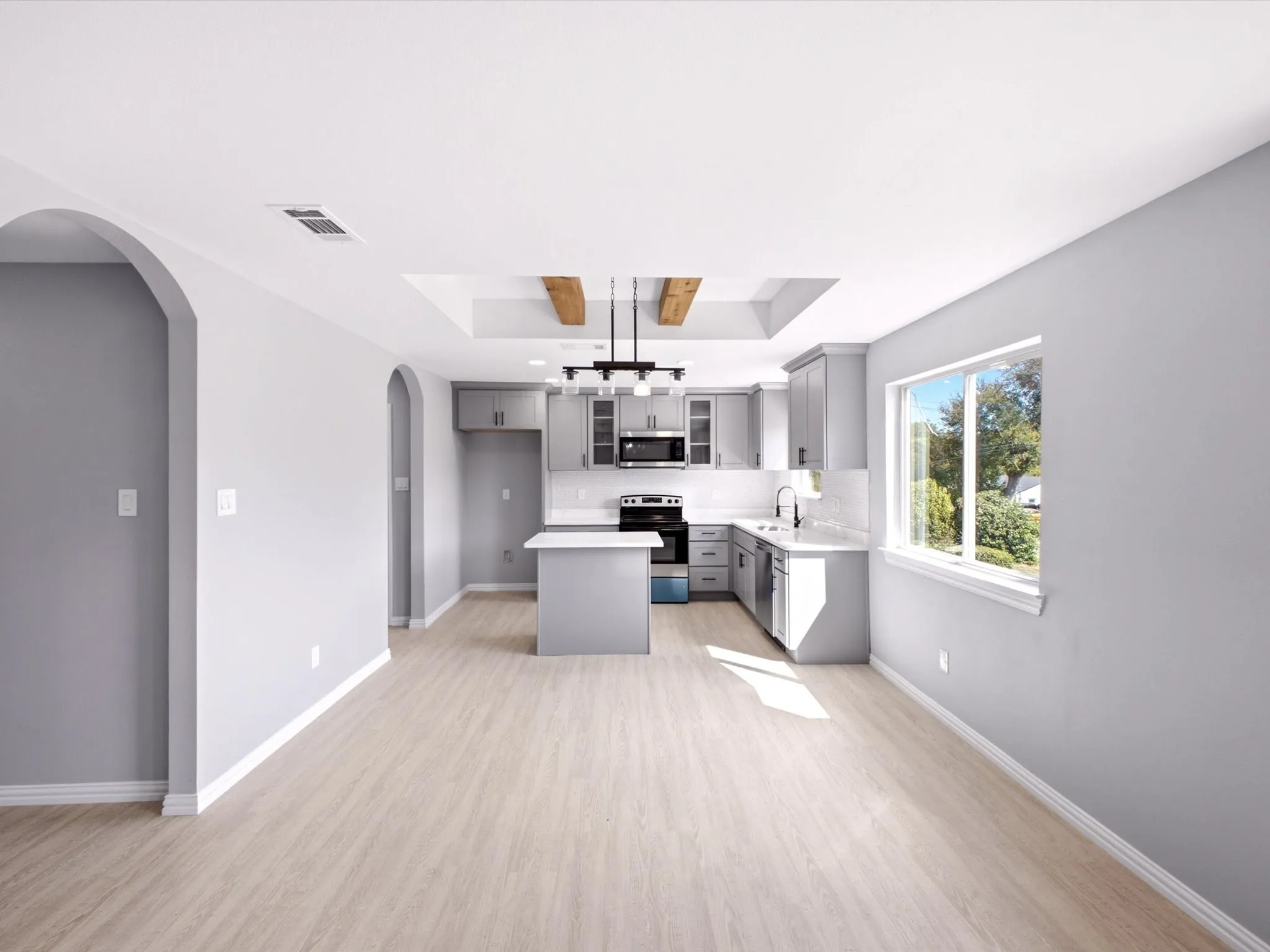 Light wood-finish flooring, arched doorways, kitchen island with seating, recessed ceiling feature with wood beams, and a modern kitchen with gray cabinetry