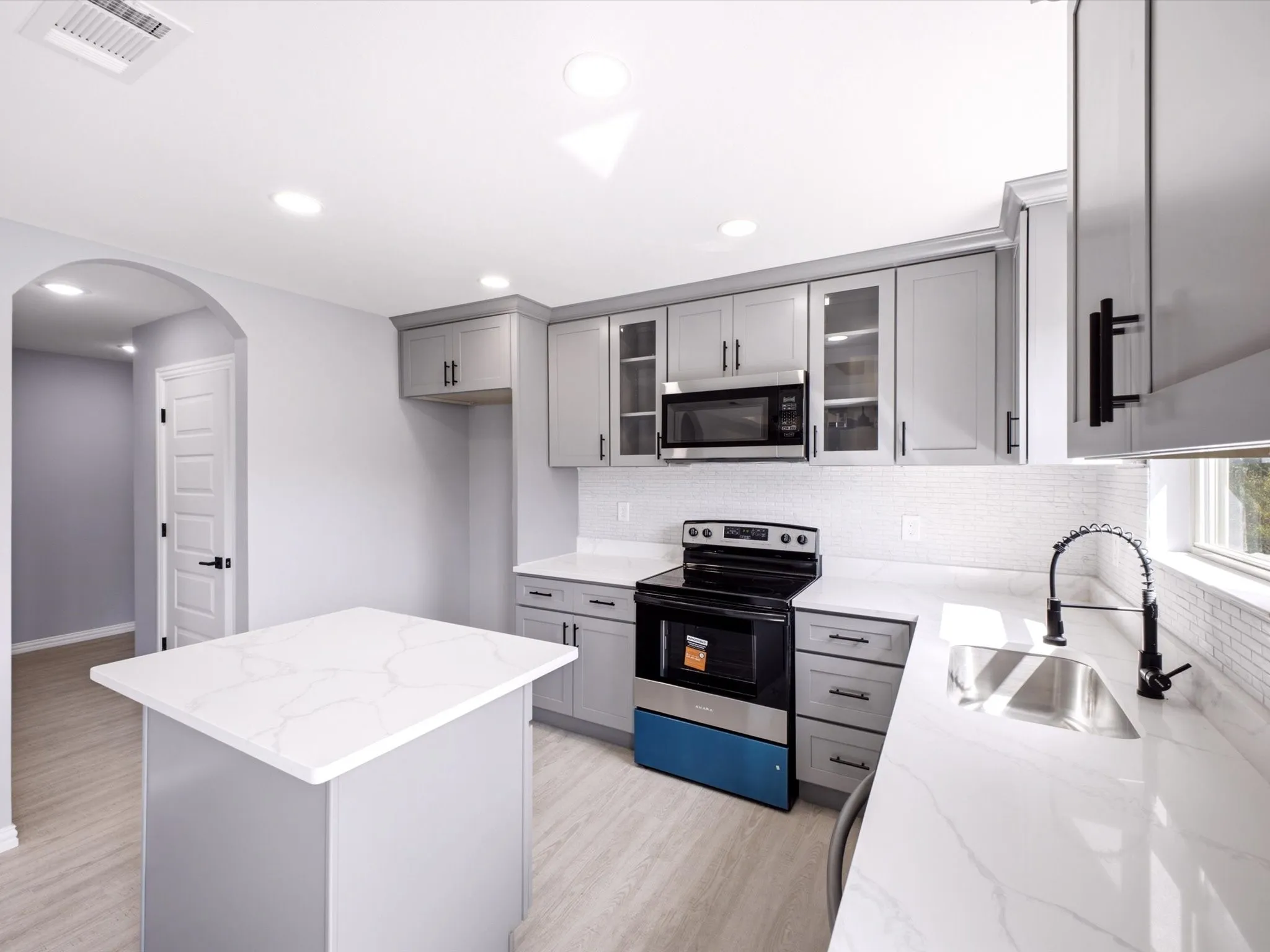 Kitchen featuring light gray cabinetry, white countertops, tile backsplash, stainless steel sink with a matte black faucet, and wood-finish flooring