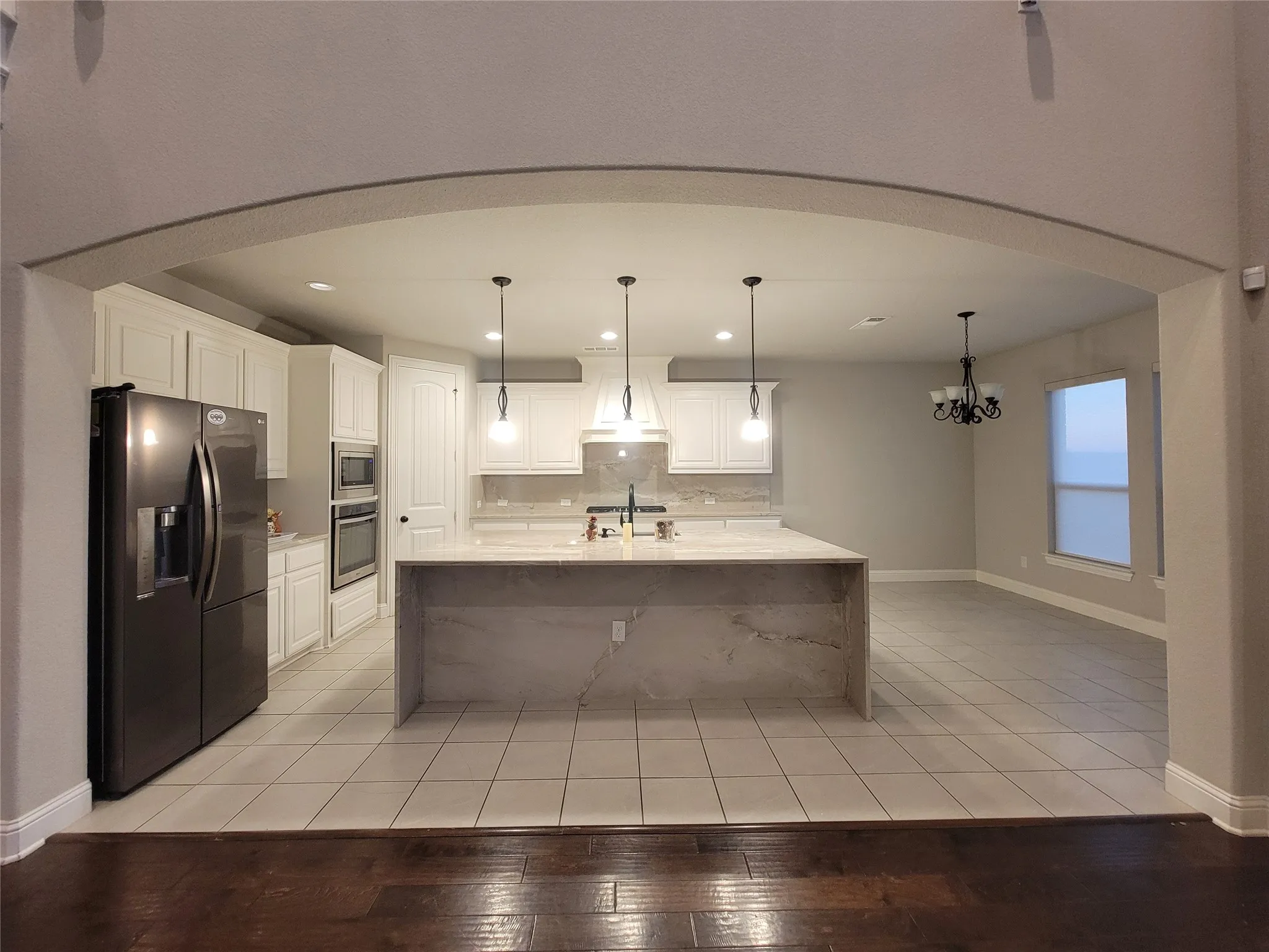 Arched entryway to a kitchen featuring an oversized island with stone-finish waterfall edge, white cabinetry, integrated double ovens, and tile flooring
