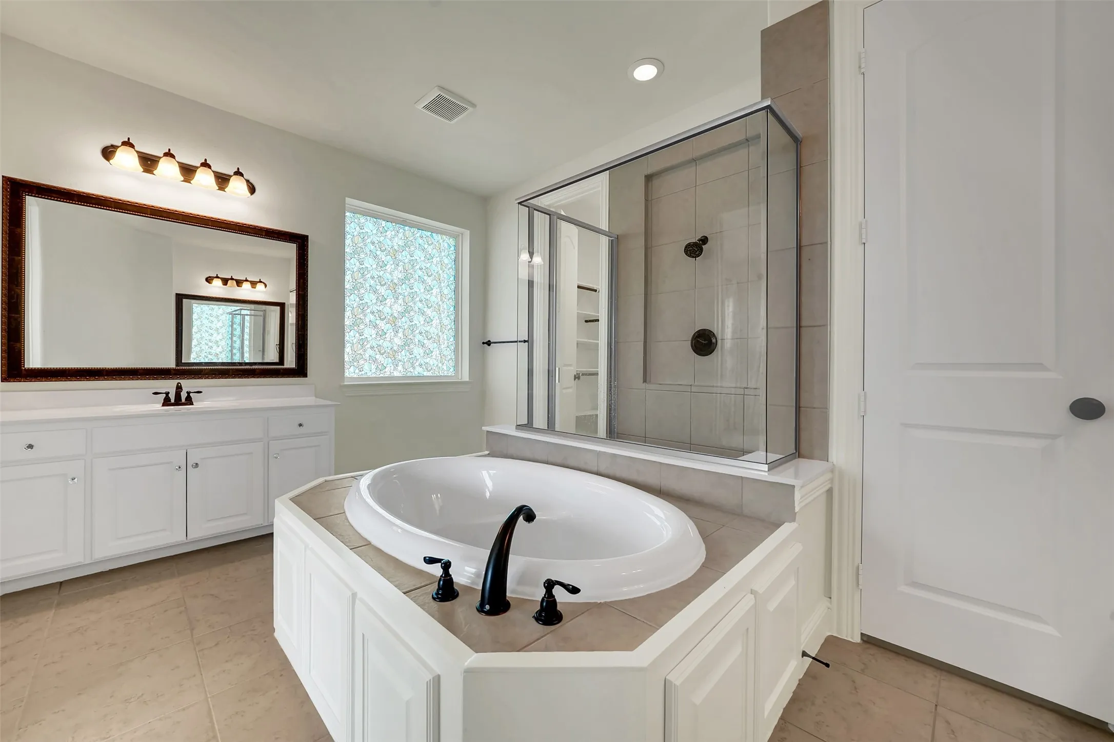 Bathroom featuring a built-in soaking tub with dark-finish fixtures, a glass-enclosed shower, a double vanity with a rectangular mirror, and light-tone floor tiling